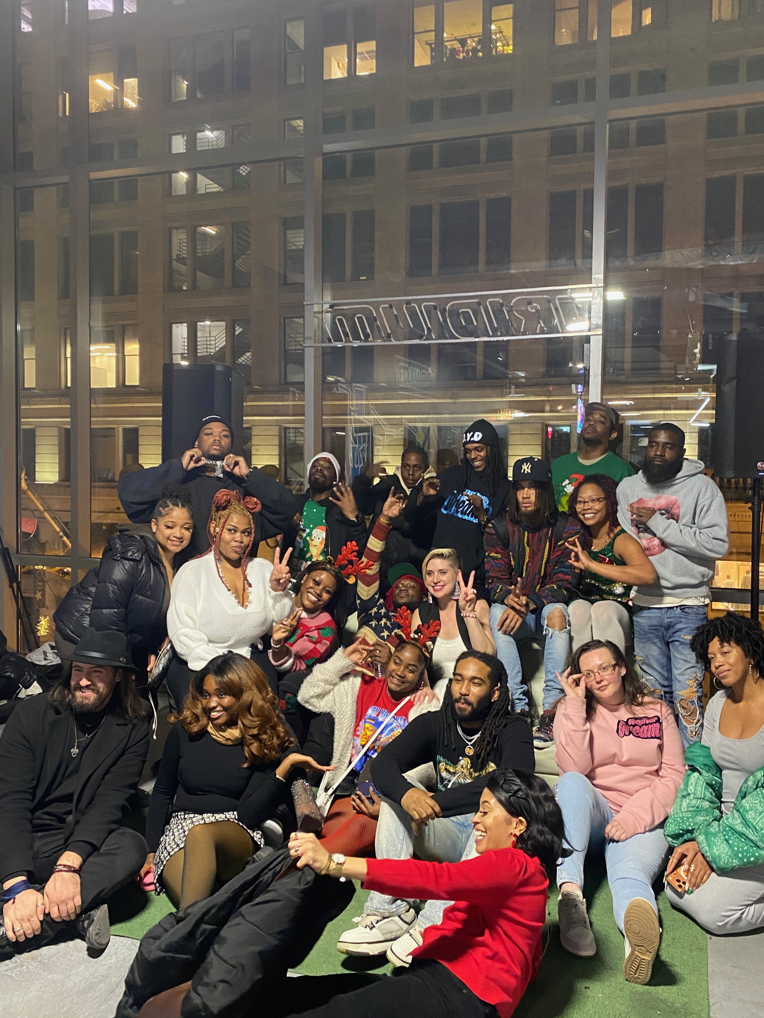 A group of diverse people, including men and women, some wearing Christmas-themed sweaters, are sitting and kneeling together inside a building with large glass windows. They are smiling, posing, and making peace signs. The background shows a reflection of a cityscape at night through the glass windows.