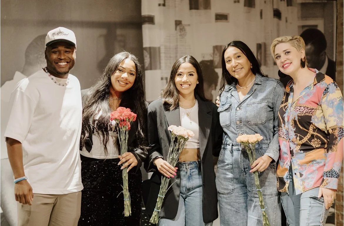 Group of five diverse people standing together, smiling, and holding flowers, inside a room with a black and white photo mural in the background.