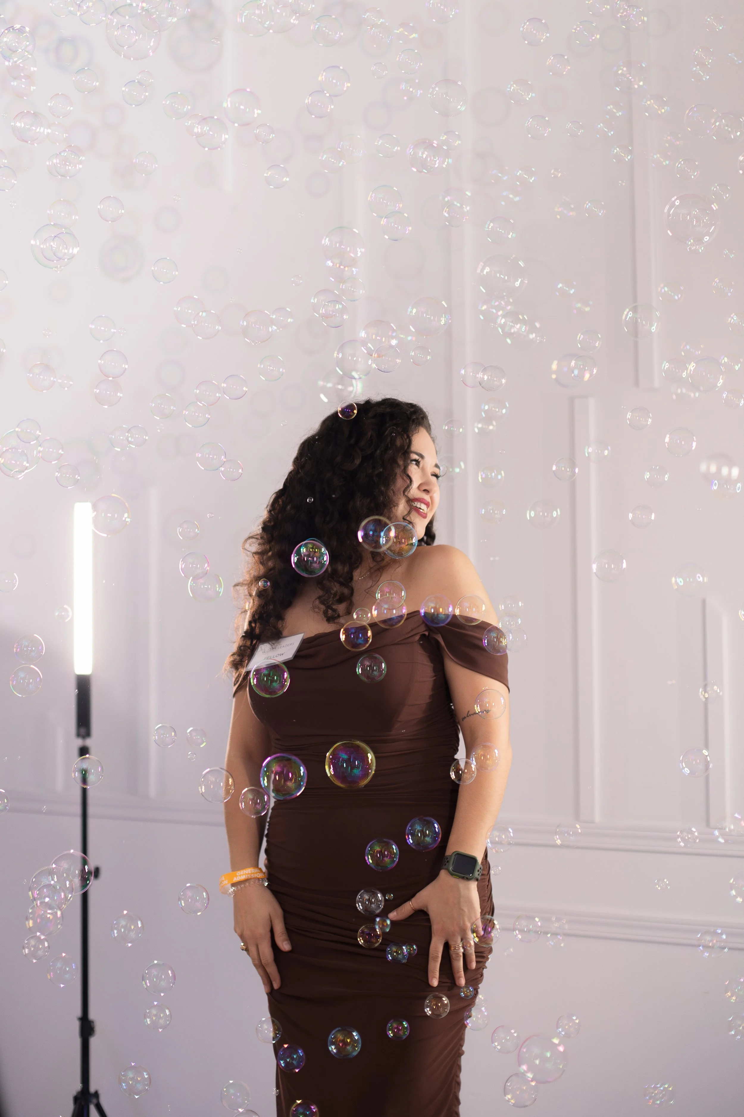 A woman with dark, curly hair wearing a fitted, off-the-shoulder brown dress stands in a room with white walls, surrounded by soap bubbles. She is smiling, looking to her left, with her right hand resting on her thigh and her left arm slightly bent. She is wearing a watch and orange wristbands on her right wrist.