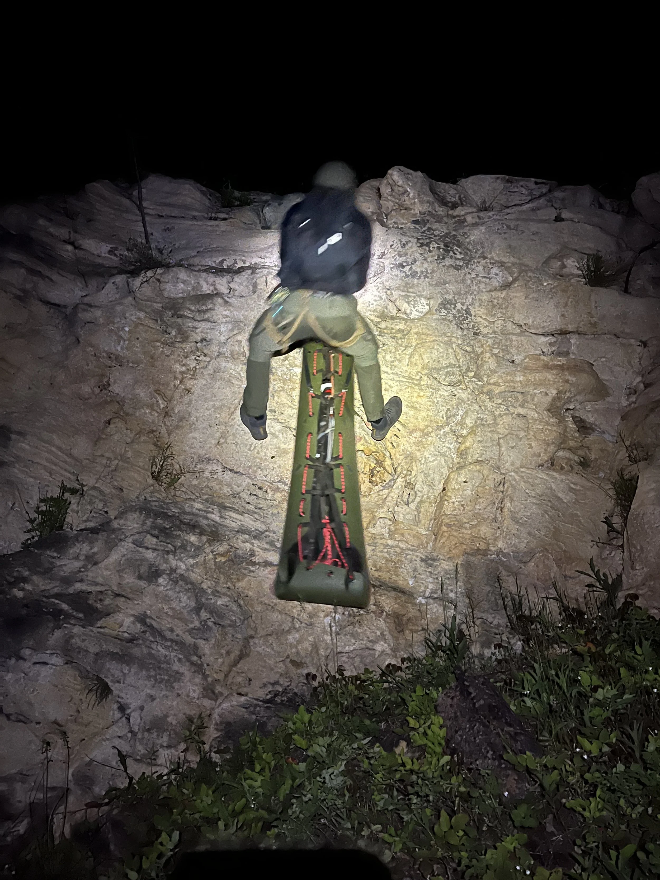 Person climbing a steep rock wall at night with a headlamp for illumination, wearing a backpack and outdoor gear.