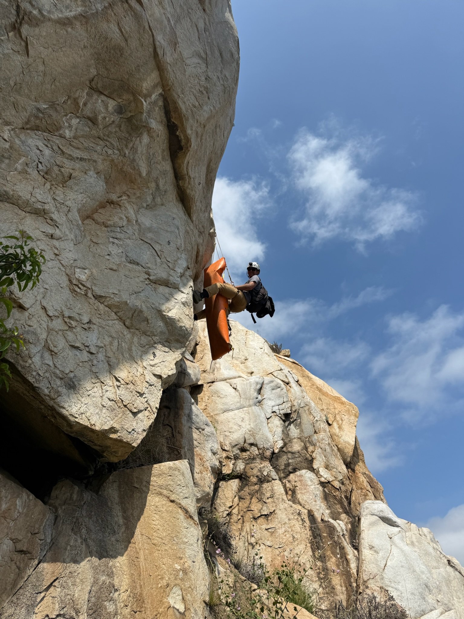 A climber ascending a tall, rugged rock face outdoors against a blue sky with a few clouds, carrying a backpack and using climbing gear.