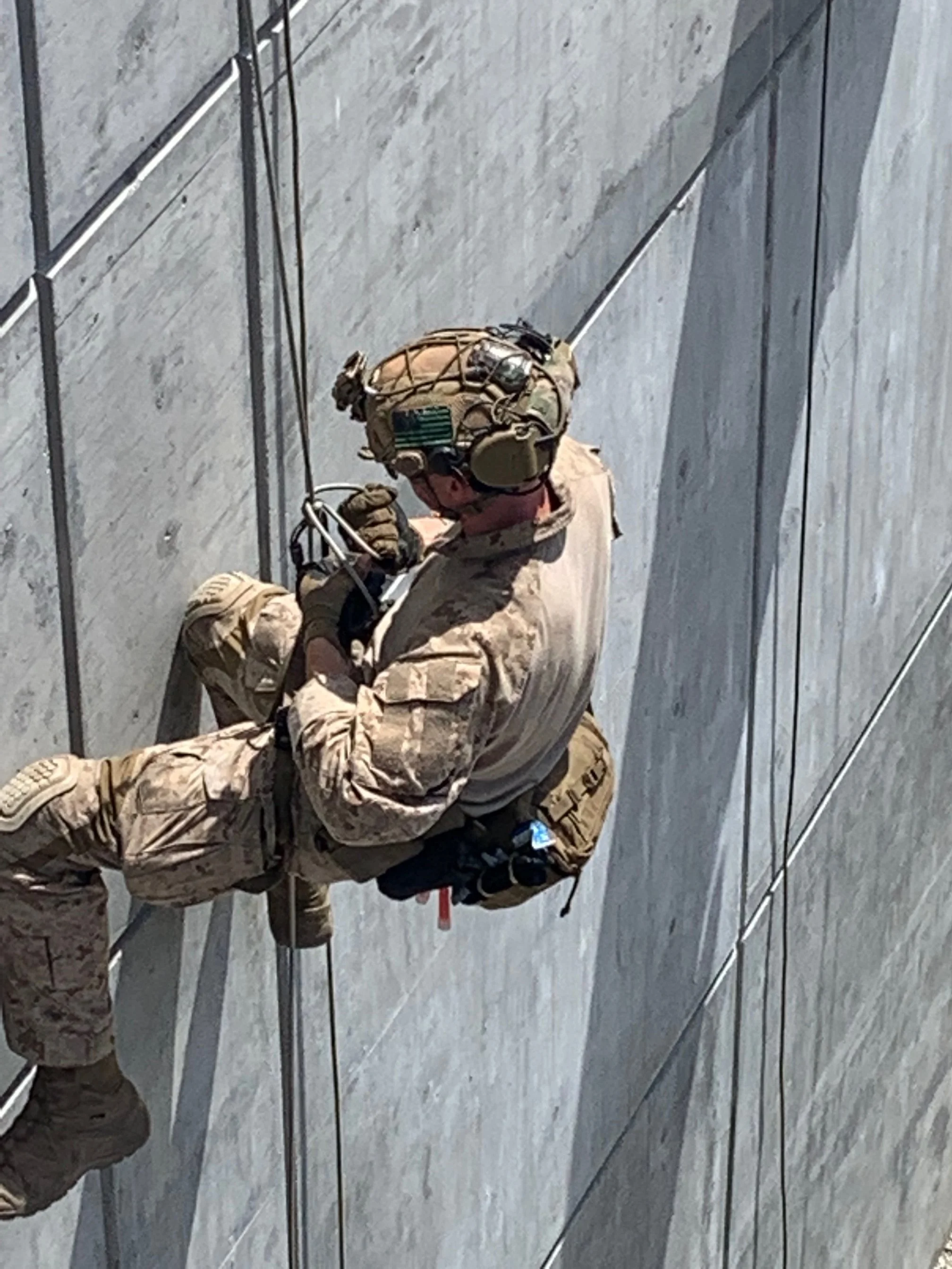 A soldier is rappelling down a concrete wall, wearing camouflage uniform and tactical gear.