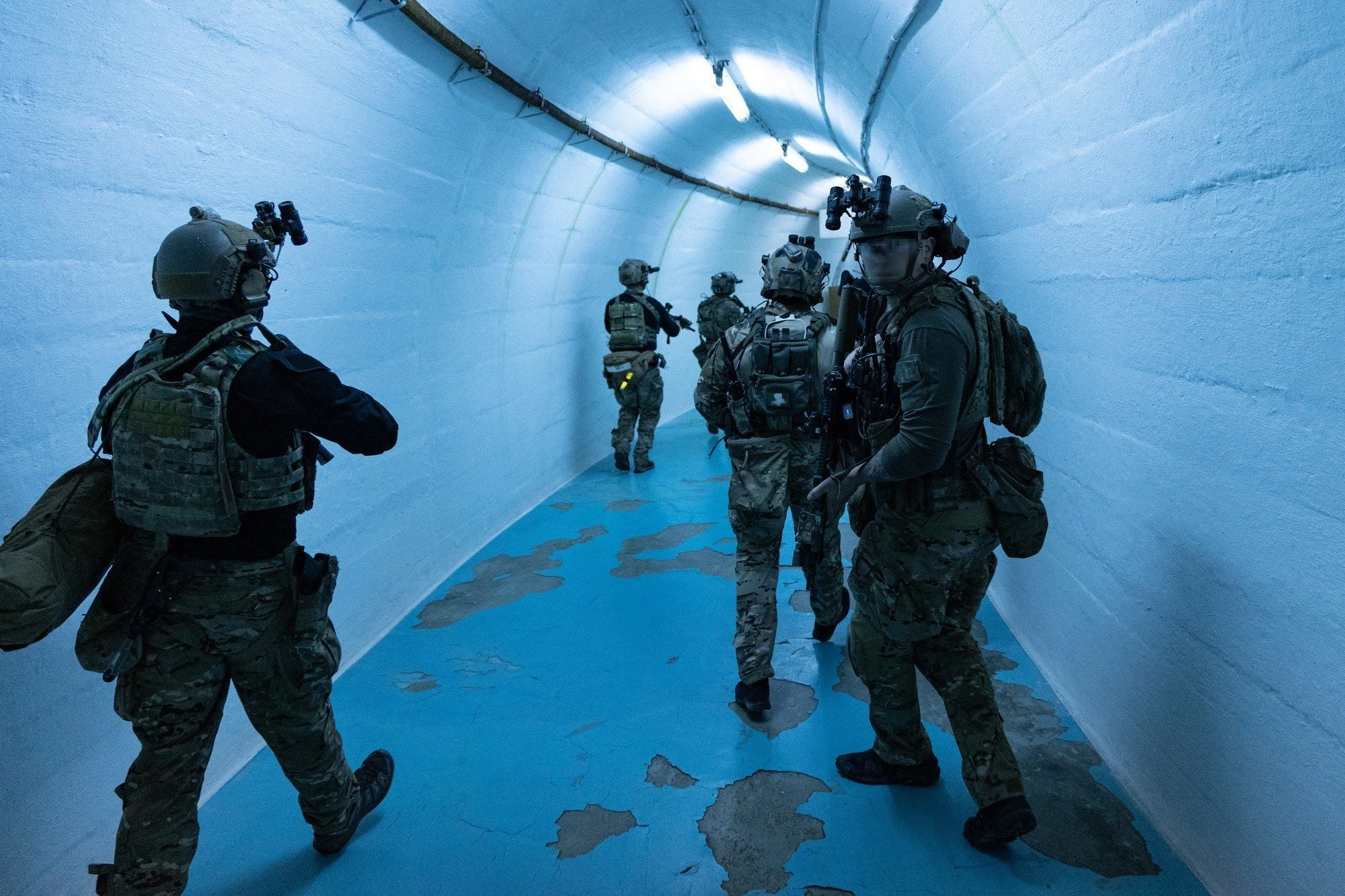 Military personnel walking through an indoor tunnel with white walls and a blue floor, wearing tactical gear and helmets.