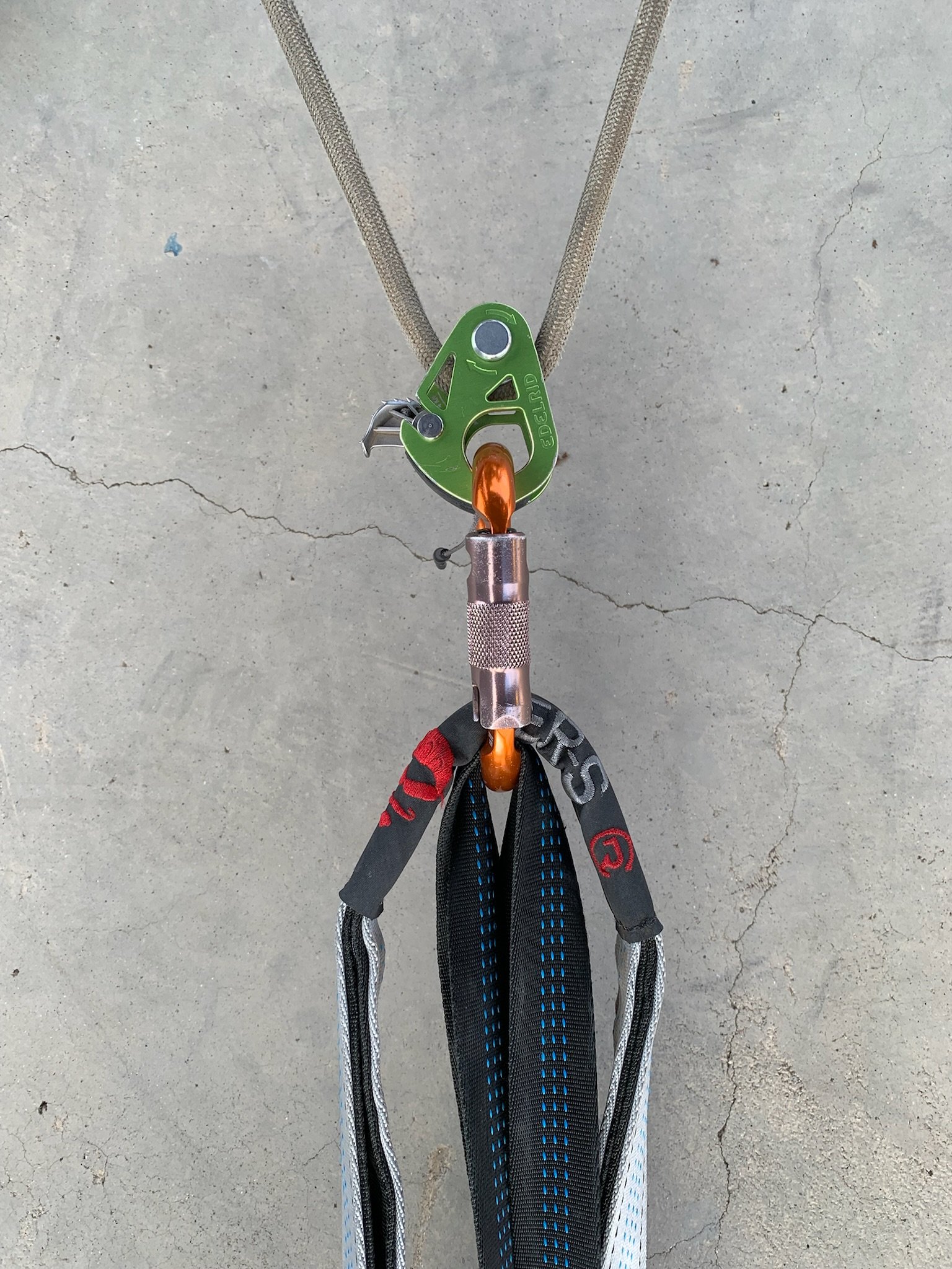 Climbing harness with a green belay device attached to an orange carabiner, black, red, blue, and gray straps hanging from the setup, on a concrete floor.