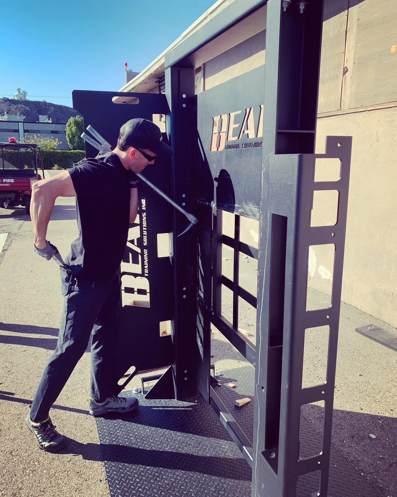 A man wearing sunglasses, a black cap, black T-shirt, and gray pants nailing the frame of a large black workout or training equipment outside on a sunny day.