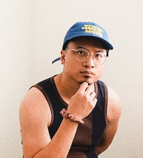 Brown Filipino-American author, miguel santiago flores, wearing glasses, a blue "Workers Deserve More" baseball cap, and a sleeveless black shirt.