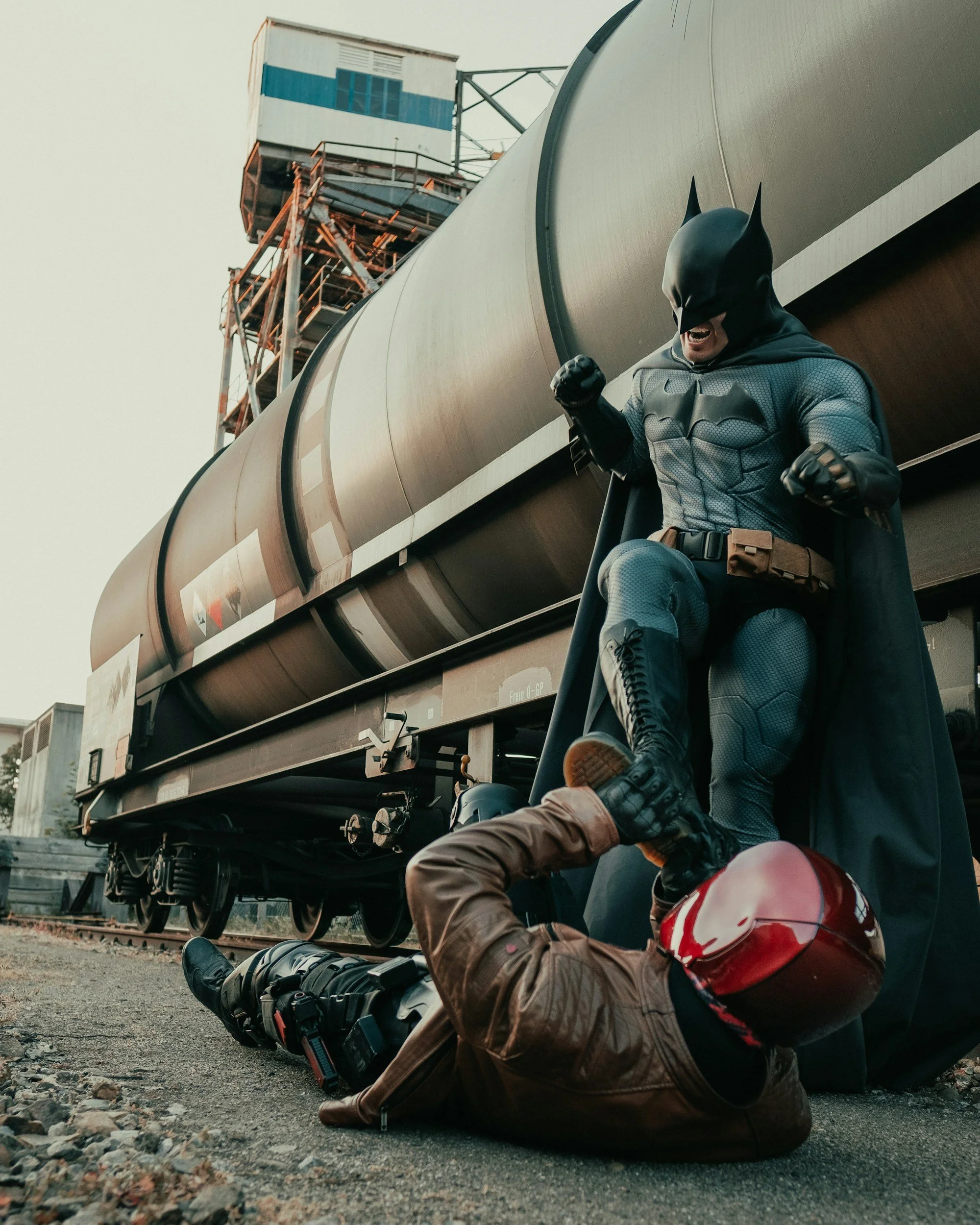 A person dressed as Batman is striking a triumphant pose with clenched fists, standing over a person dressed as a police officer who is lying on the ground at a train track, with a large train and an industrial structure in the background.