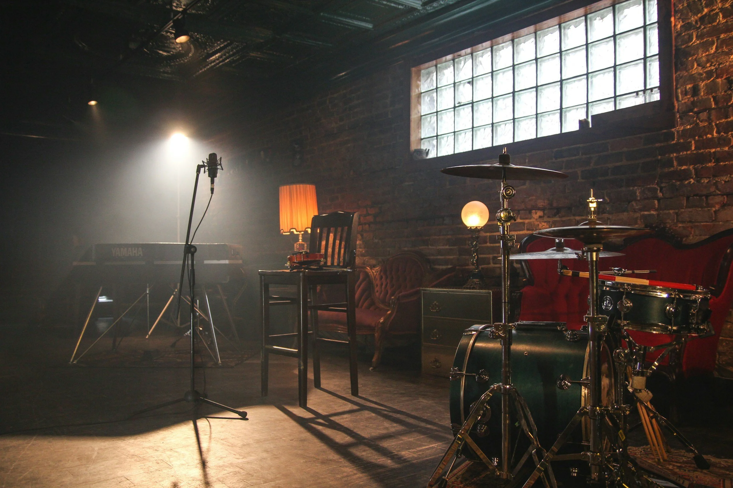 Music studio with musical instruments and vintage furniture, including a drum set, keyboard, microphone, chairs, and lamps, illuminated by warm lighting in a brick-walled room.