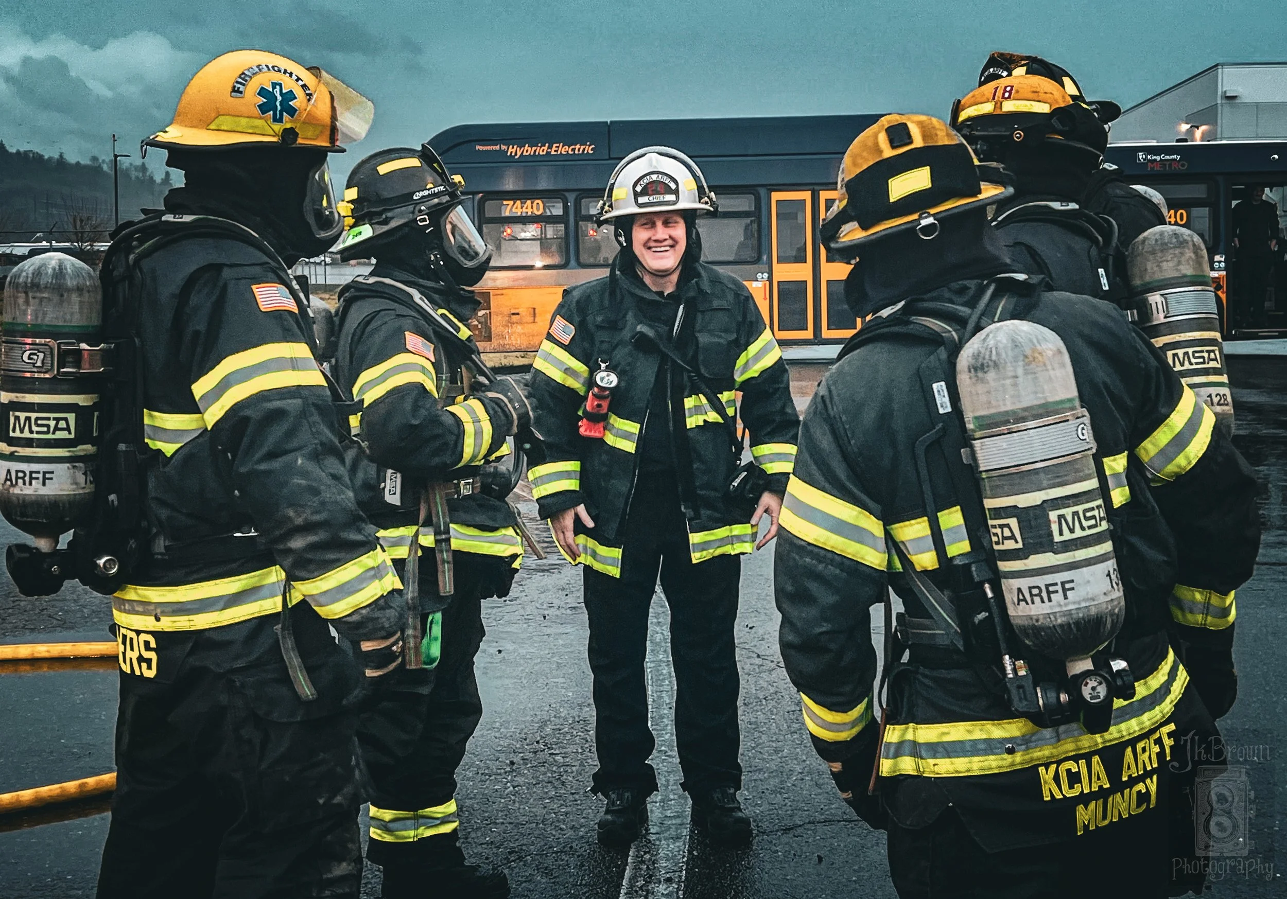 Group of firefighters in firefighting gear standing in front of a yellow bus, with an overcast sky.