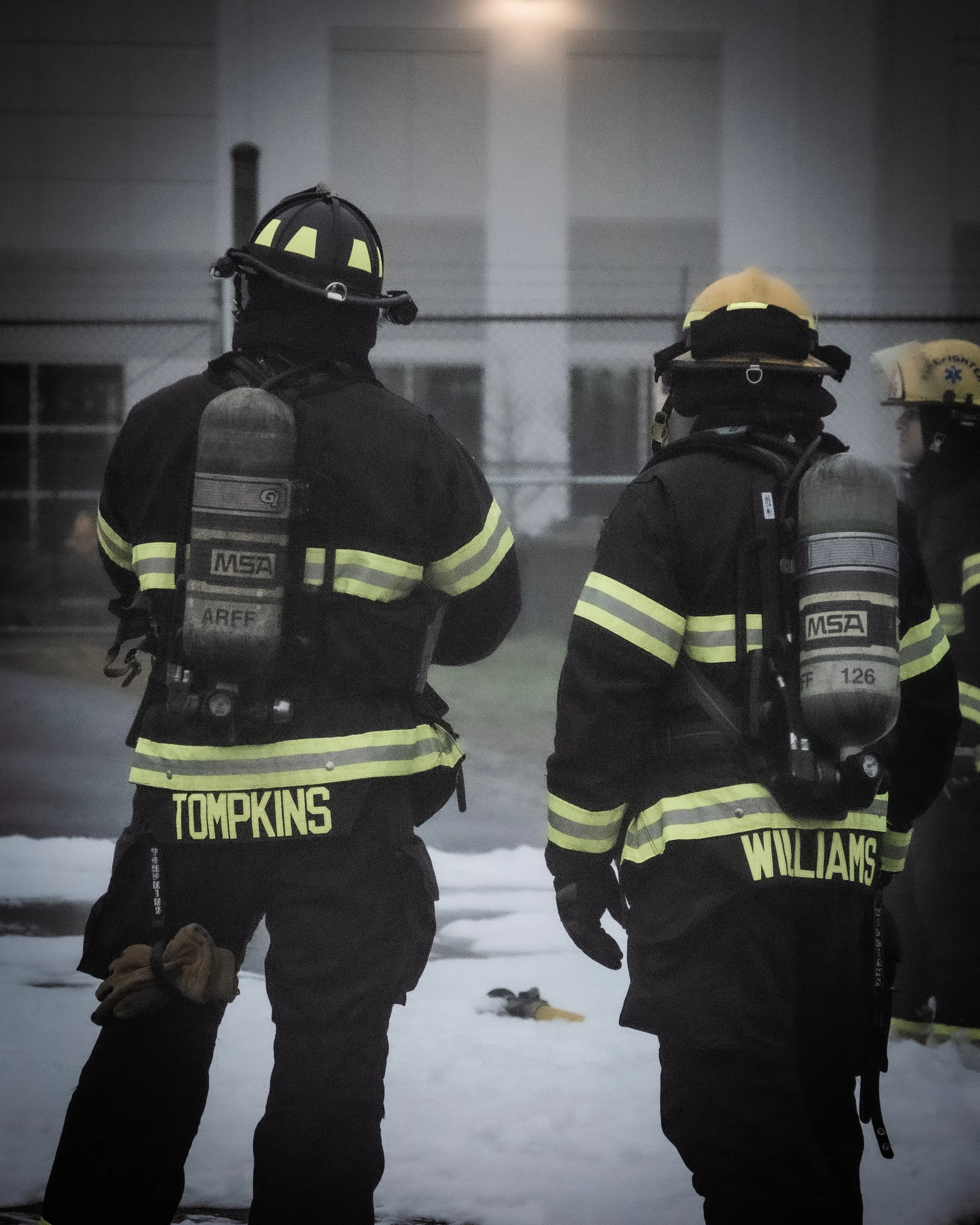 Two firefighters in full gear, including helmets and oxygen tanks, standing on snow near a building.