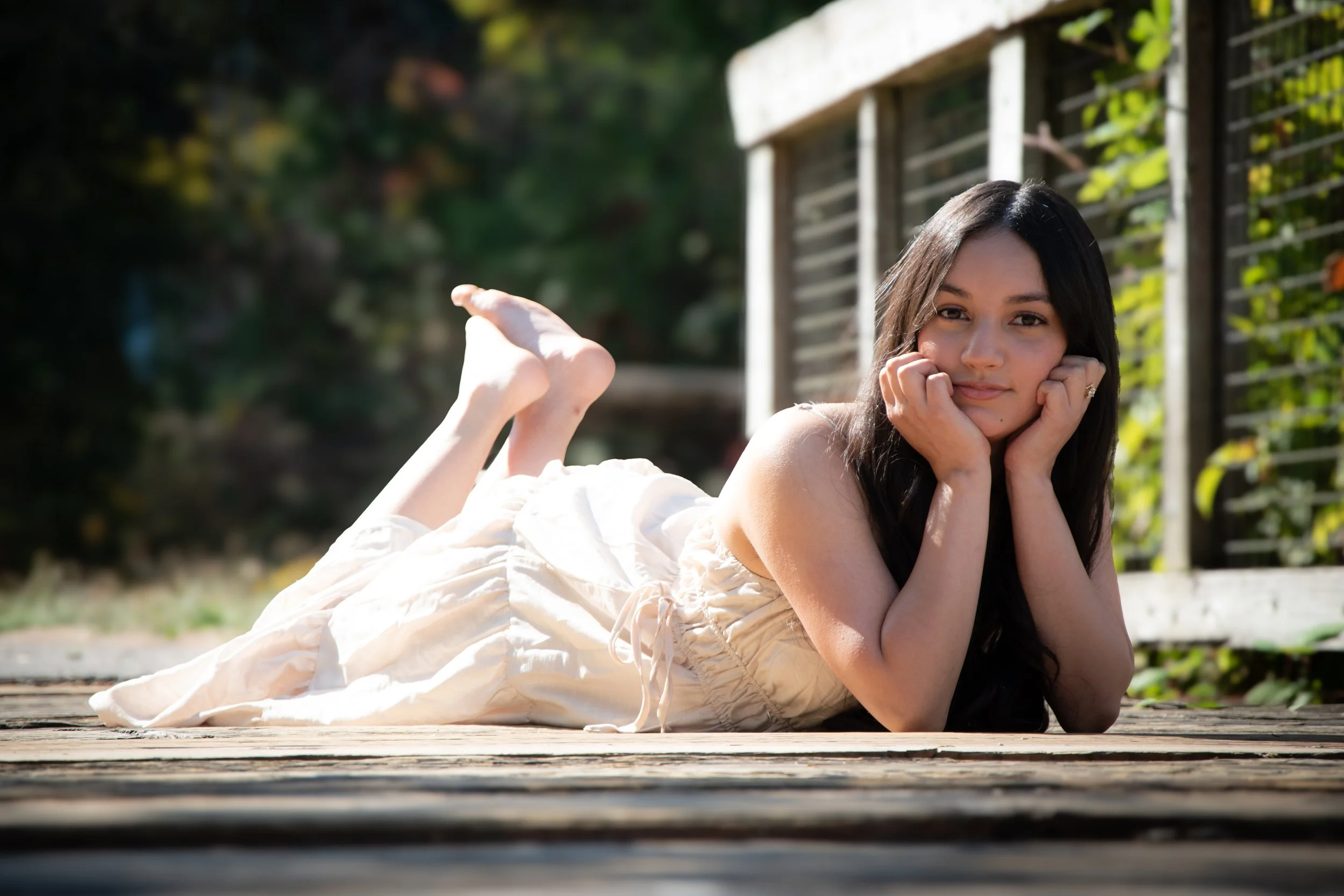 Young woman lying on a wooden deck outdoors, resting her chin on her hands, with a relaxed expression. She has long dark hair and is wearing a light-colored dress, with a garden or greenery background behind her.