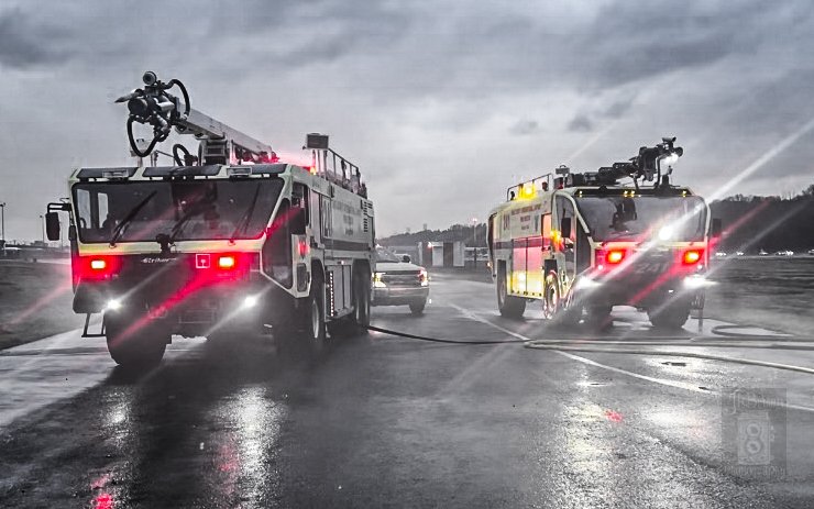 Firefighters' trucks spraying water on the wet pavement during rainy weather.