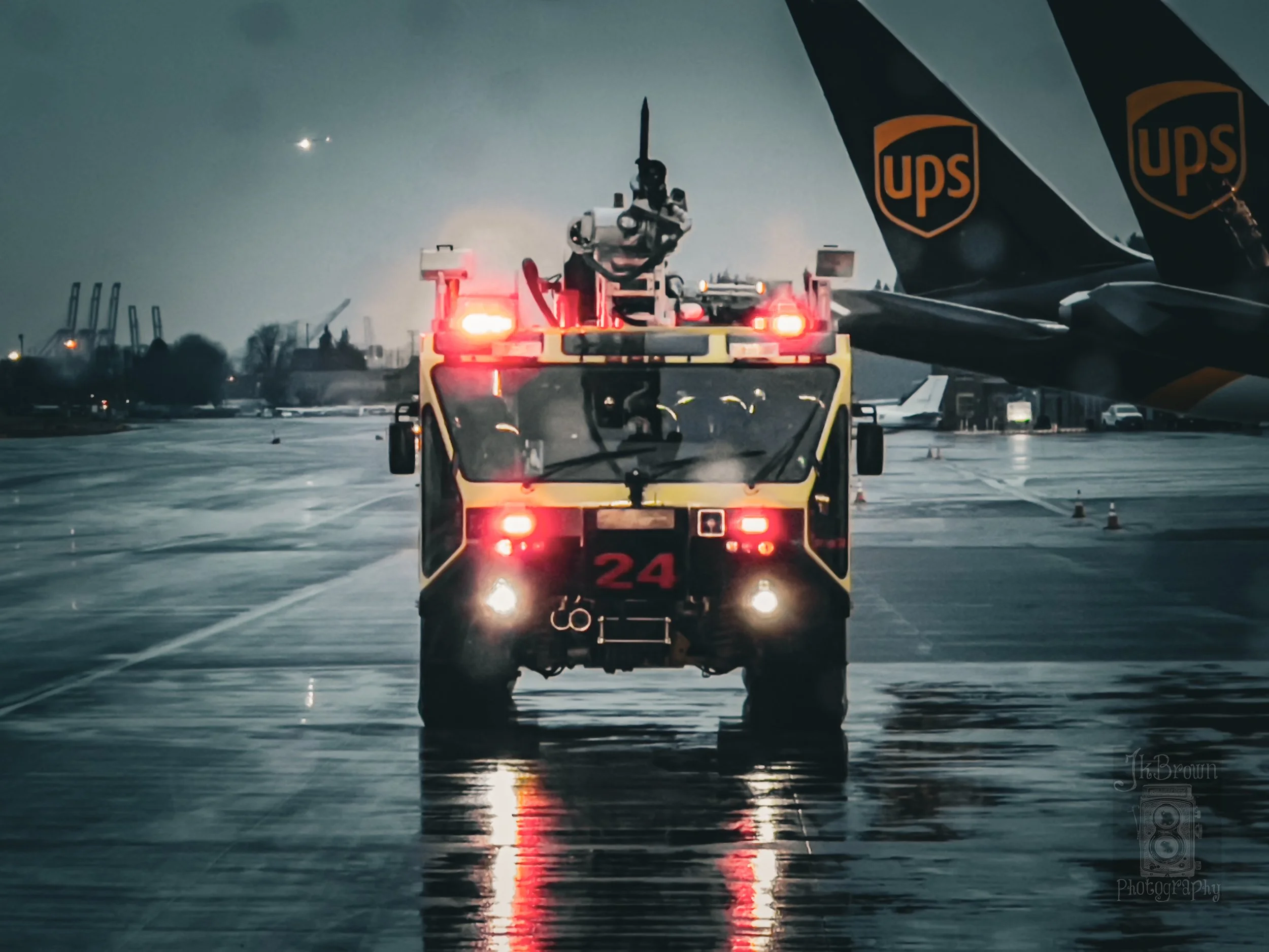 Fire truck driving on wet airport tarmac at night with an airplane wing labeled UPS in the background.