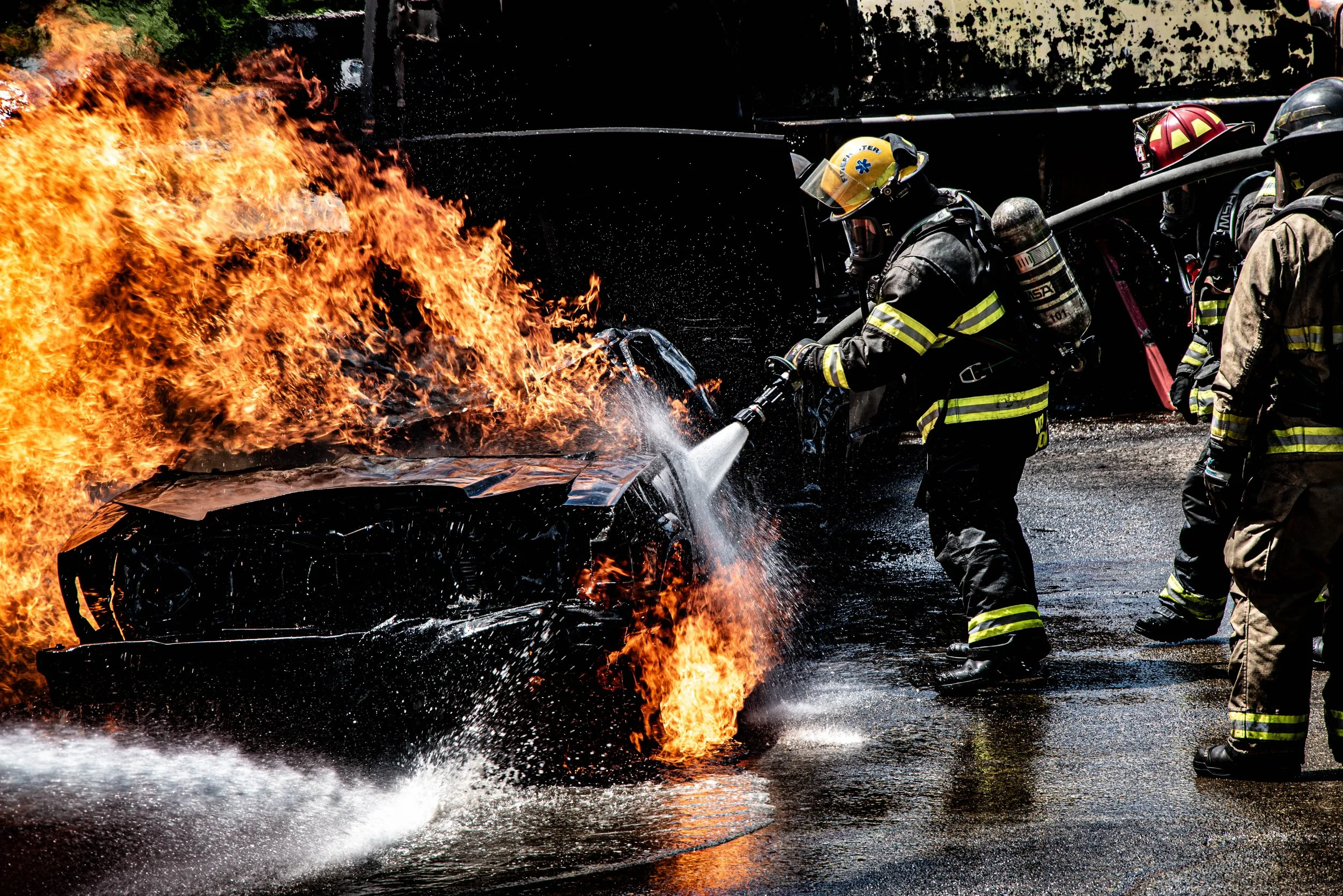 Firefighters combat a car fire, using a hose to spray water on a burning vehicle with flames and smoke.