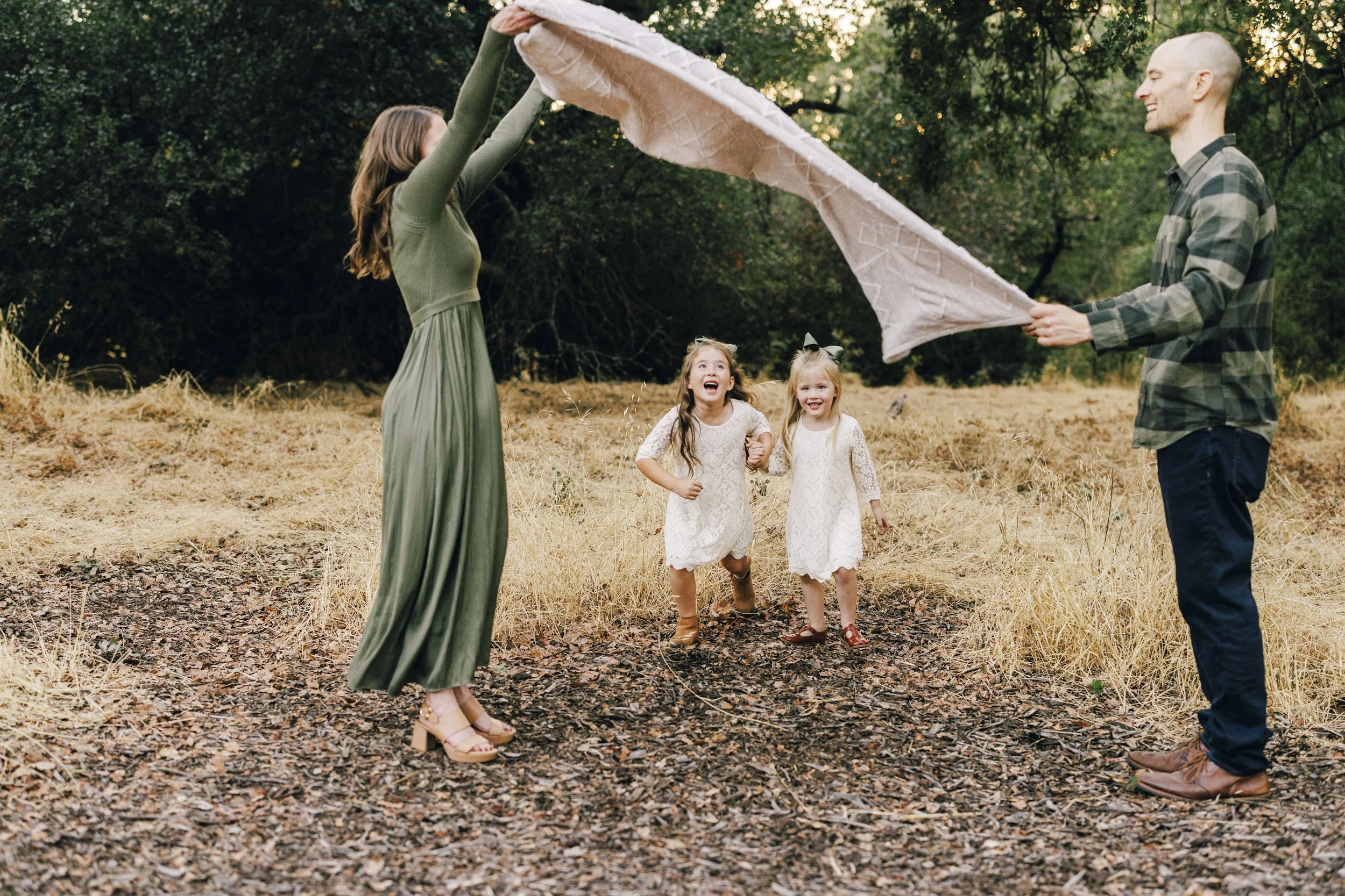 A family of four playing outdoors in a forest clearing, with two young girls in white dresses running happily while their parents lift a blanket over their heads.