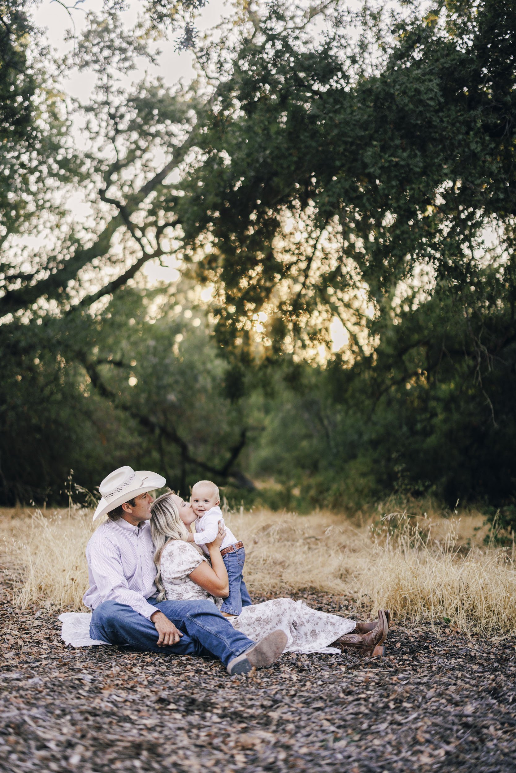 A family of three sitting on the ground in a natural outdoor setting, with trees and sunlight in the background. The man is wearing a cowboy hat, while the woman is kissing their baby on the cheek. The baby is smiling. They are dressed in casual, cou