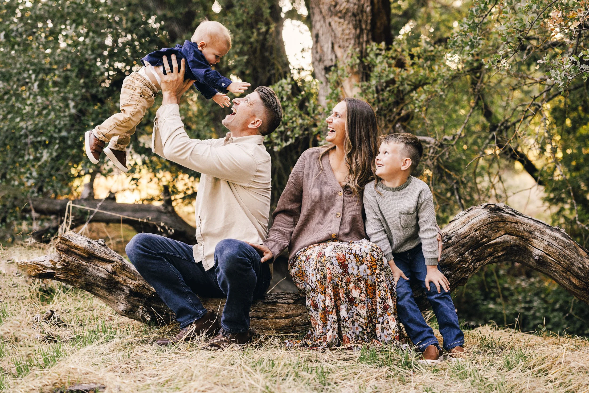 Family of four outdoors in a wooded area, sitting on a fallen tree; the father lifts a young child into the air while the mother and older son watch and smile.