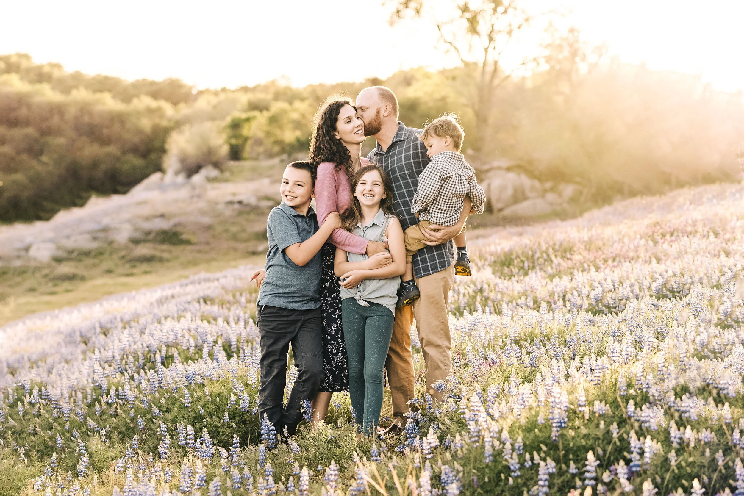 A family of six standing together in a field of lupines near Folsom Lake during sunset, sharing a loving moment with the father kissing the mother on the cheek and the children smiling.