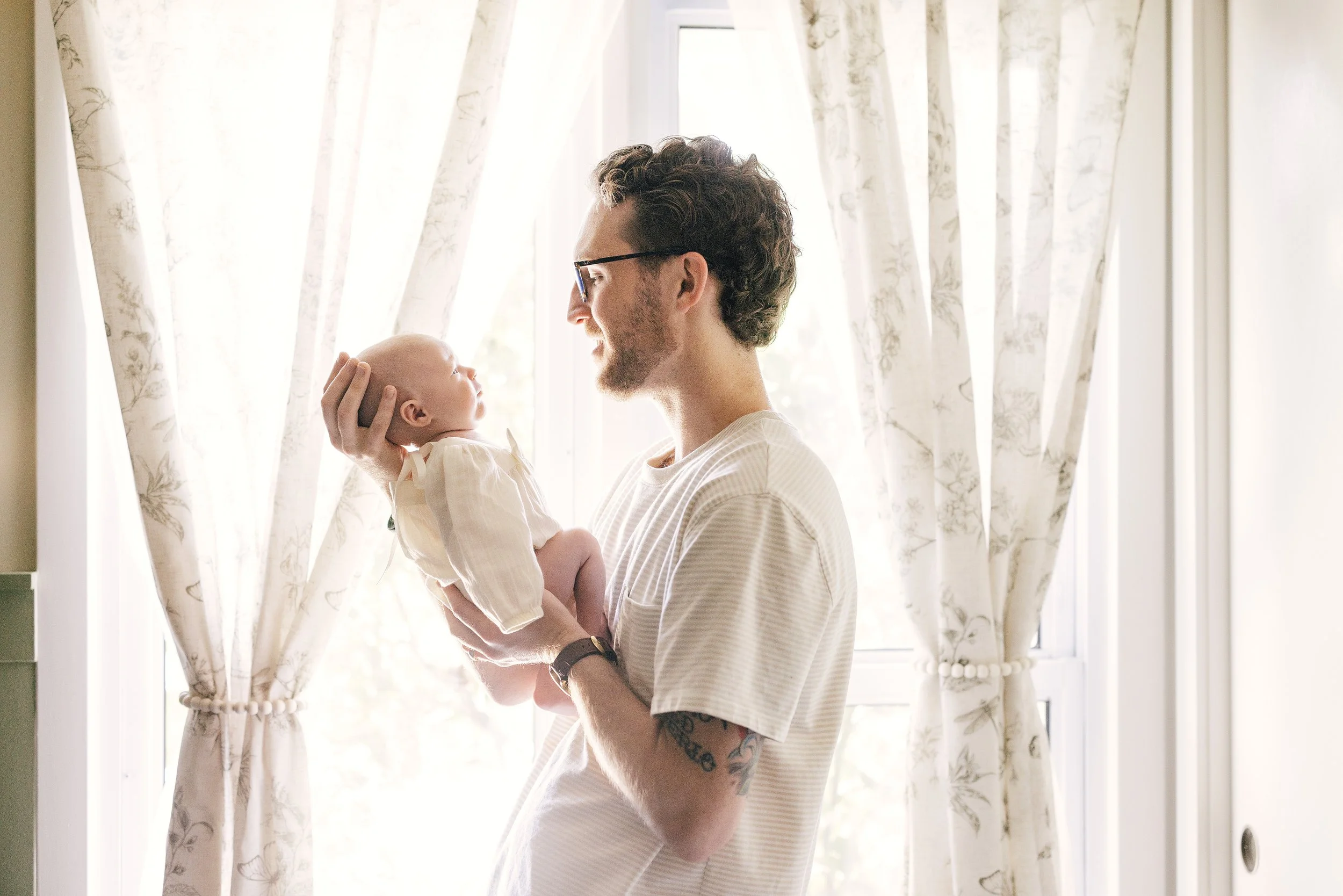 Man holding a baby inside near a window with floral curtains.