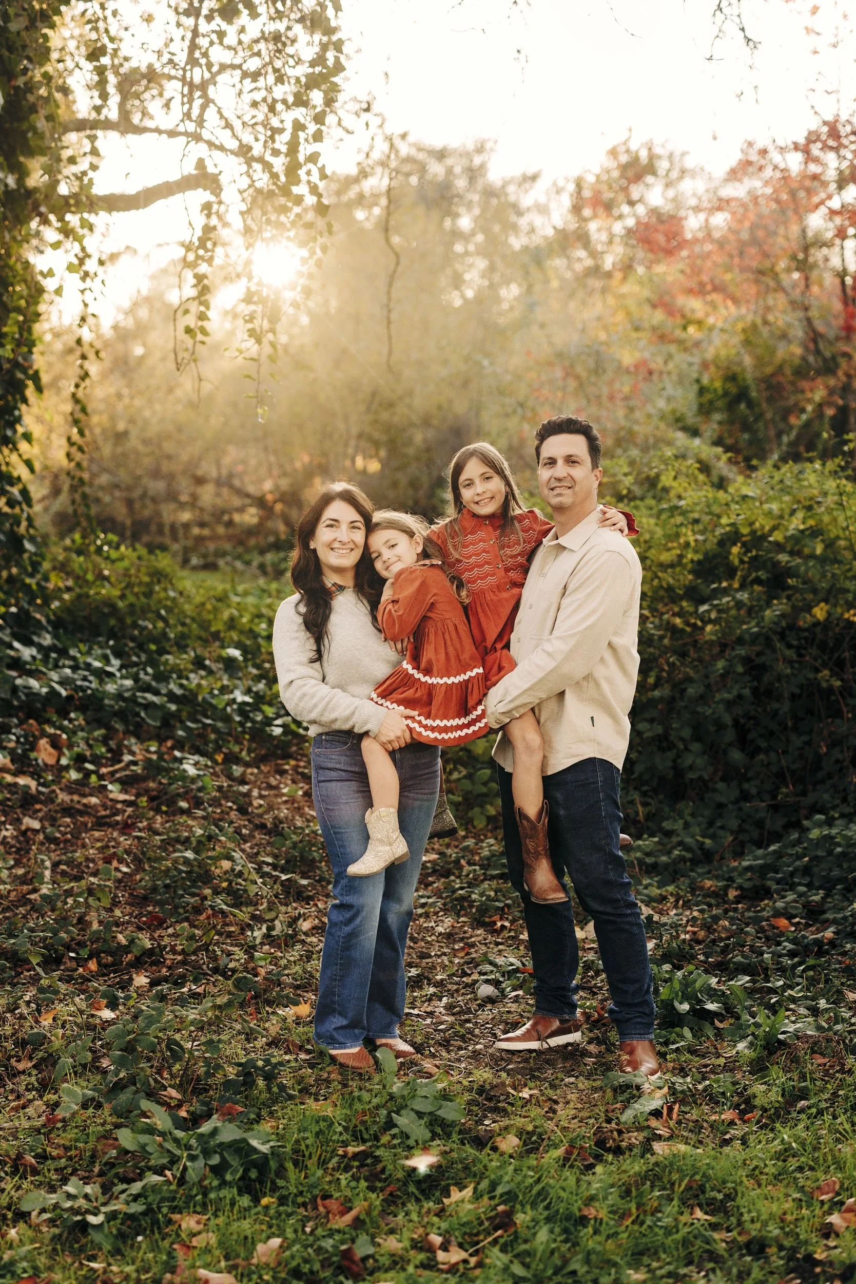 A family of four standing outdoors in a forest at sunset, smiling and holding each other. The family includes a father, mother, and two daughters, with the girls wearing matching red dresses.