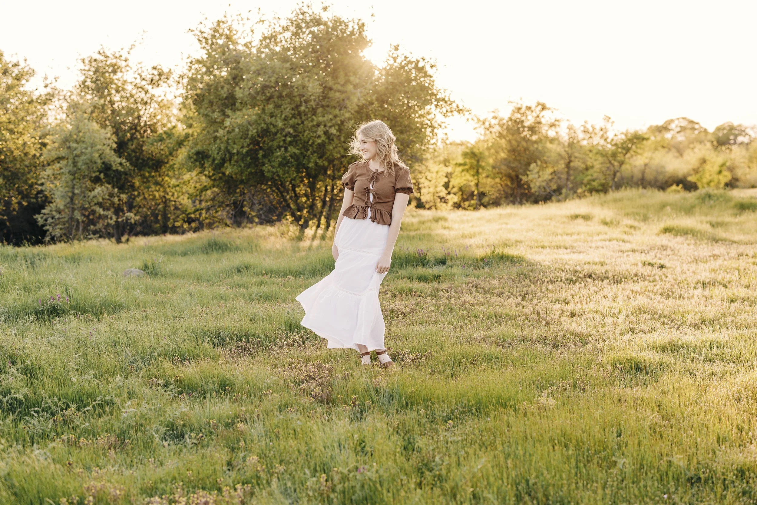 A woman with blonde hair in a white dress and brown top walking through a grassy field at sunset, with trees in the background.