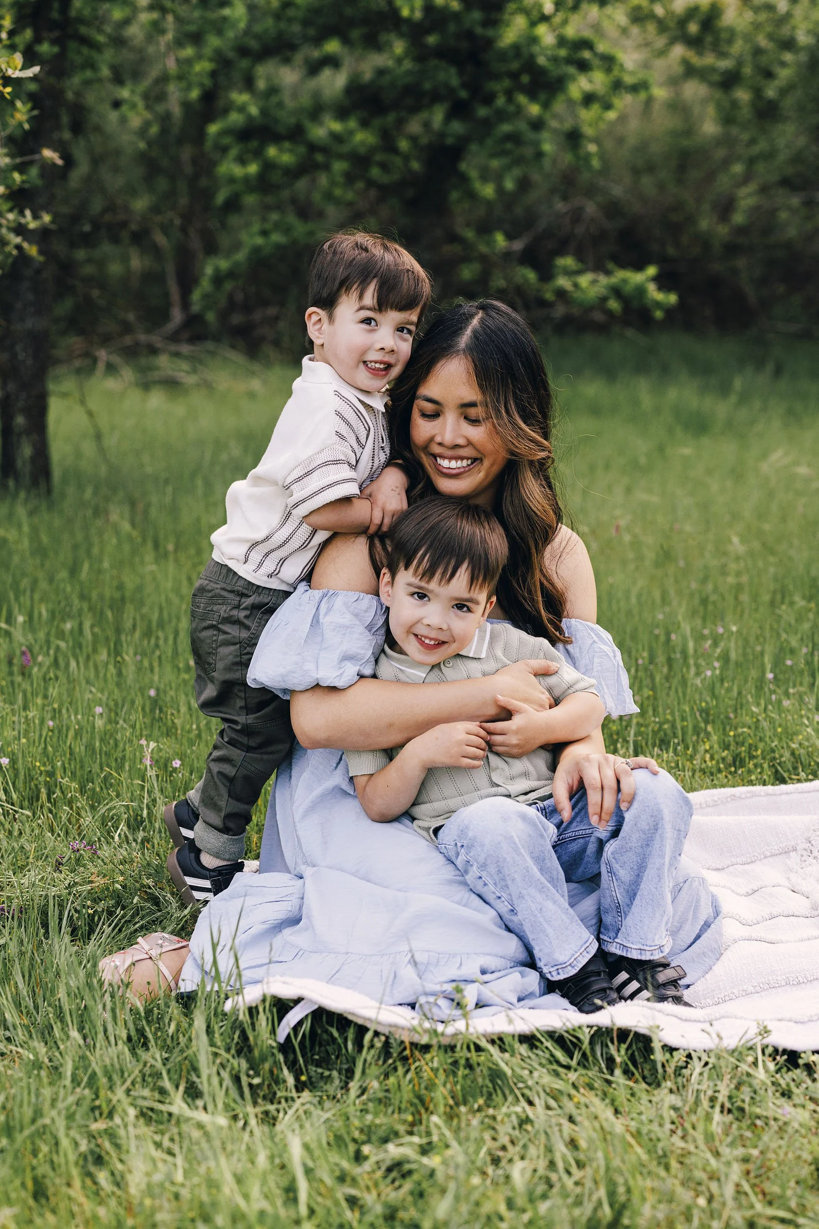A woman sitting on a blanket in a grassy field with two young boys, all smiling and enjoying nature.