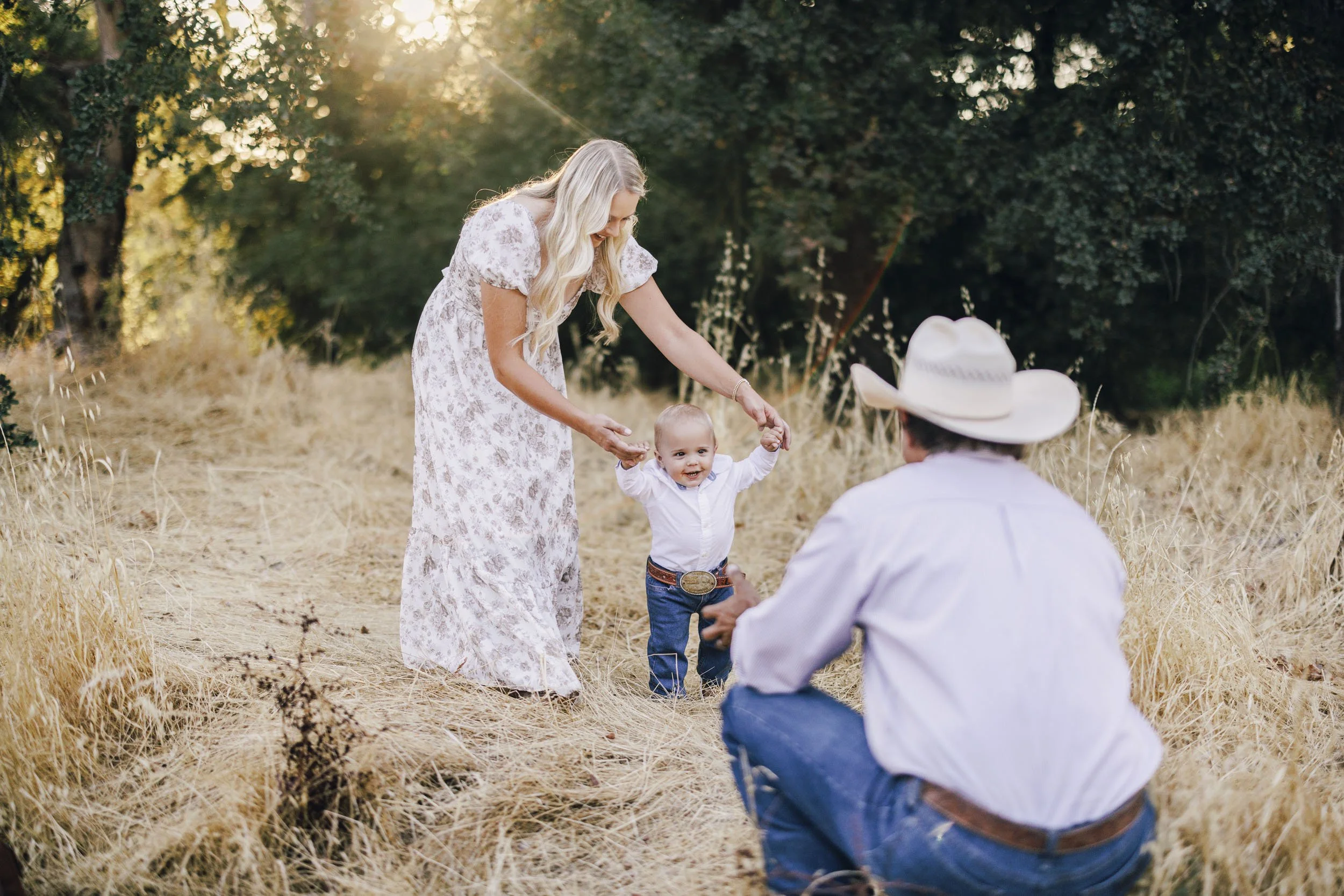 A woman in a floral dress is holding a smiling toddler by the hands while a man in a cowboy hat and white shirt kneels in front of them. They are outdoors in a field with tall dried grass and trees in the background, during late afternoon sunlight.