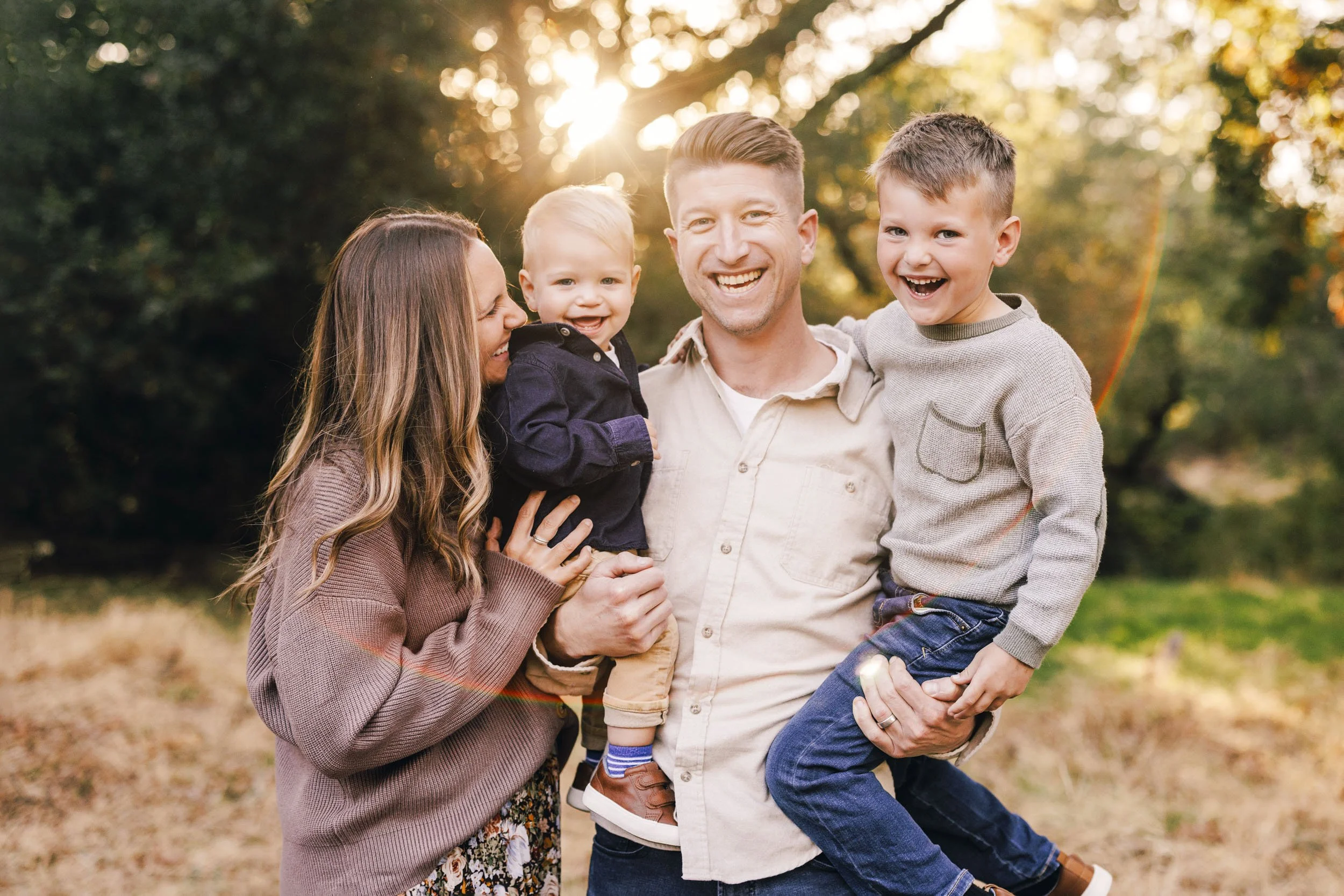 A happy family of five outdoors during sunset, with trees and foliage in the background. The family members are smiling and appear to be enjoying each other's company.