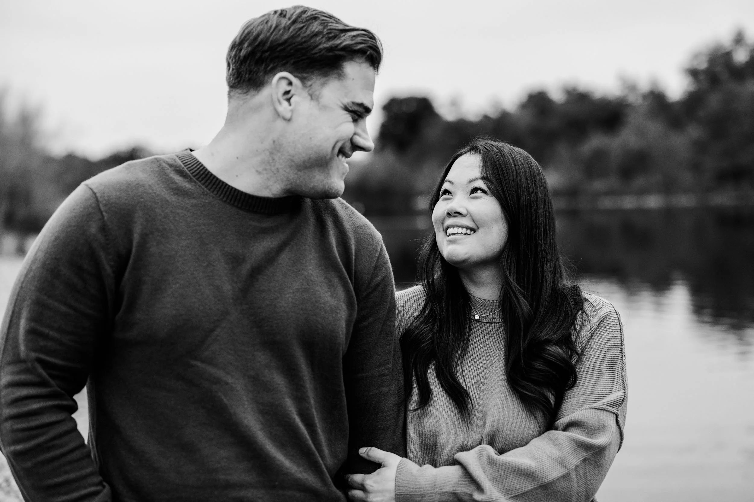 A smiling man and woman standing outdoors near a body of water, looking at each other affectionately, with trees blurred in the background.