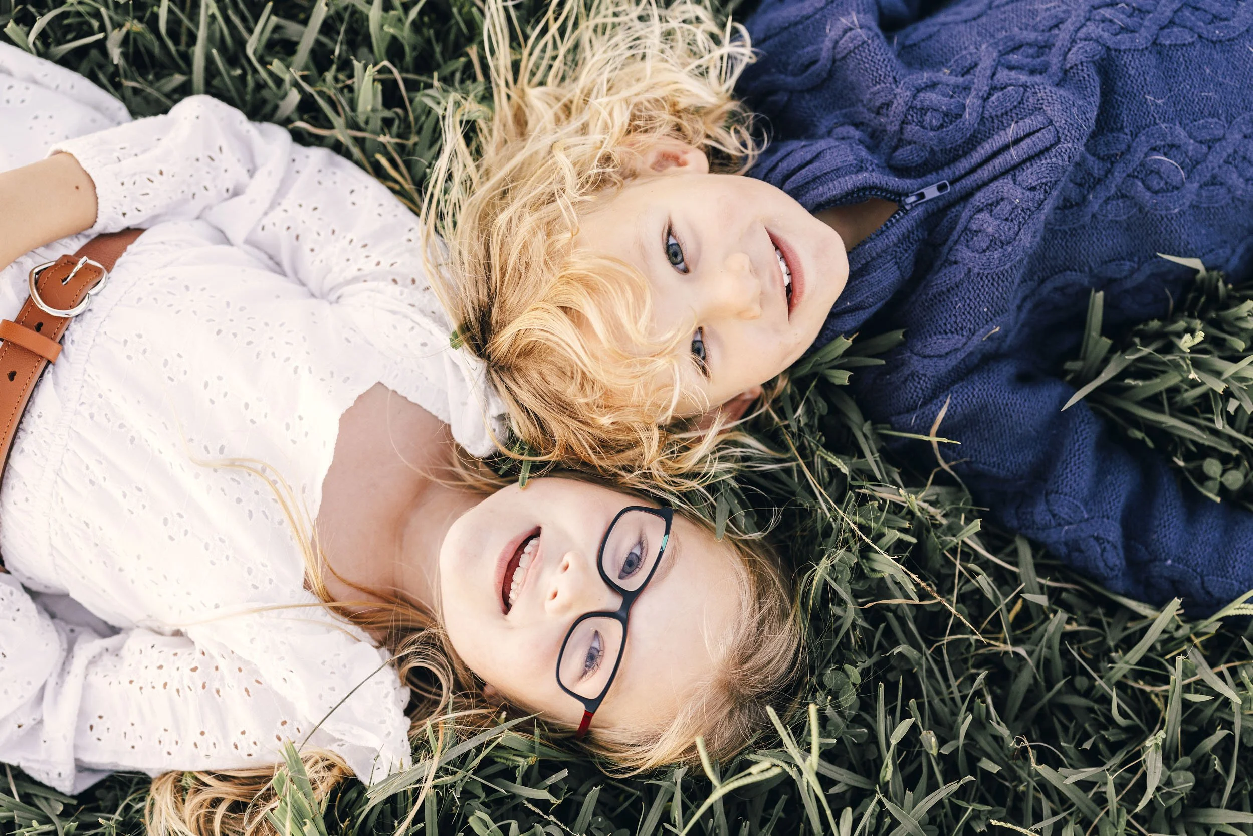 Siblings lying on grass, one with blonde curly hair, wearing a white eyelet dress, and the other with blonde straight hair, wearing glasses and a blue sweater, smiling at the camera.