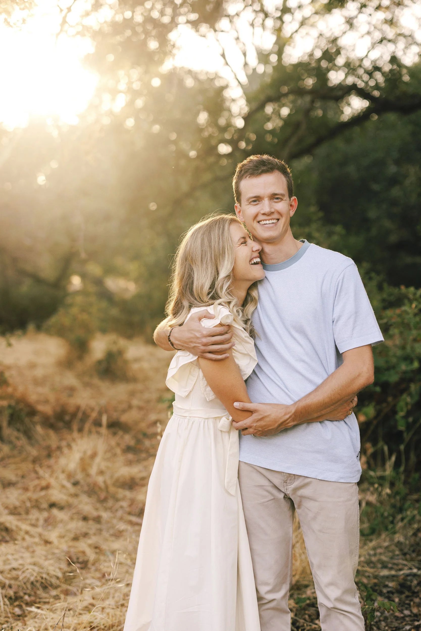 A smiling couple is hugging outdoors during sunset, with trees and a grassy area in the background.