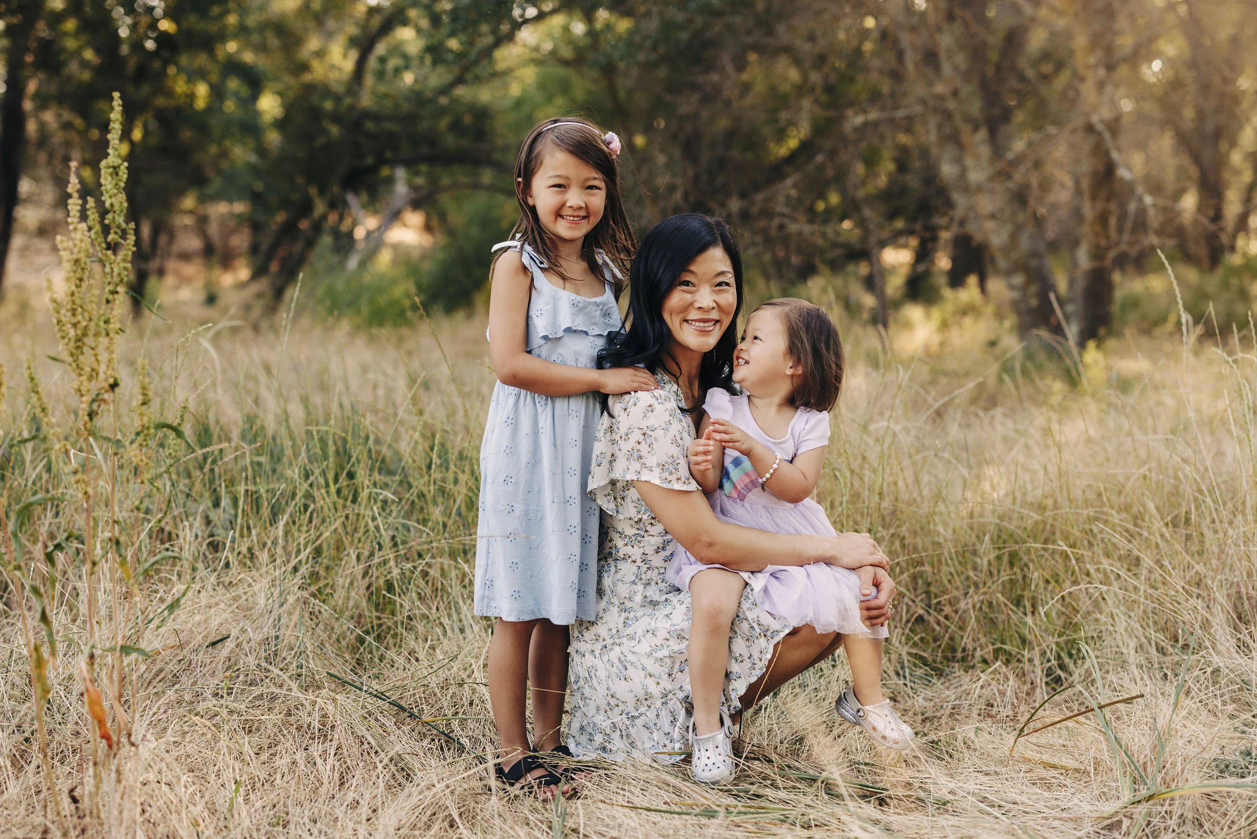A smiling woman crouches in tall grass with two young girls, one standing and one sitting on her knee, in a forested outdoor setting during golden hour.