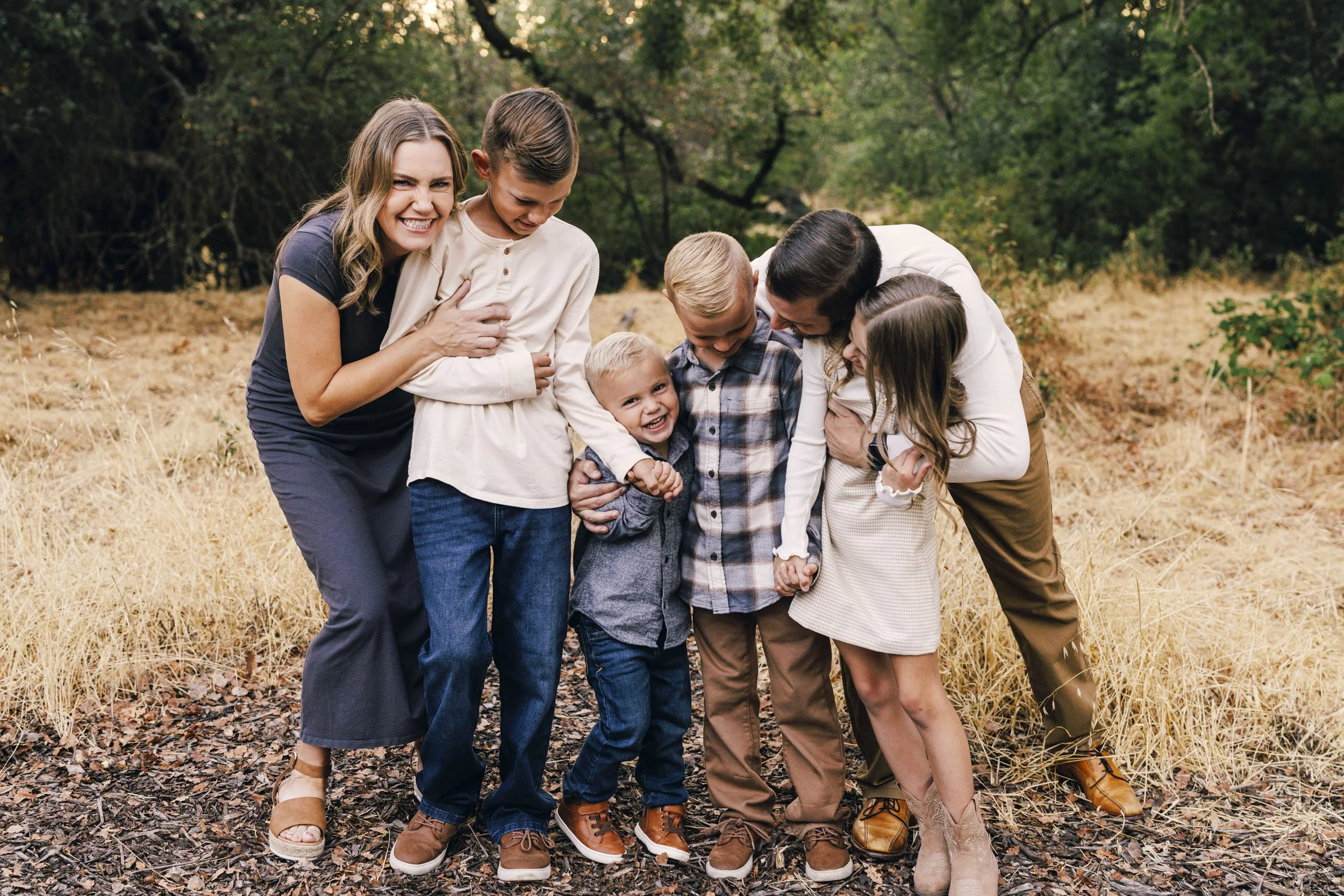 A family of six, including two adults and four children, standing outdoors in a natural setting with trees and grass, smiling and laughing together during a heartfelt photo session with Amy Wright in Roseville, California.