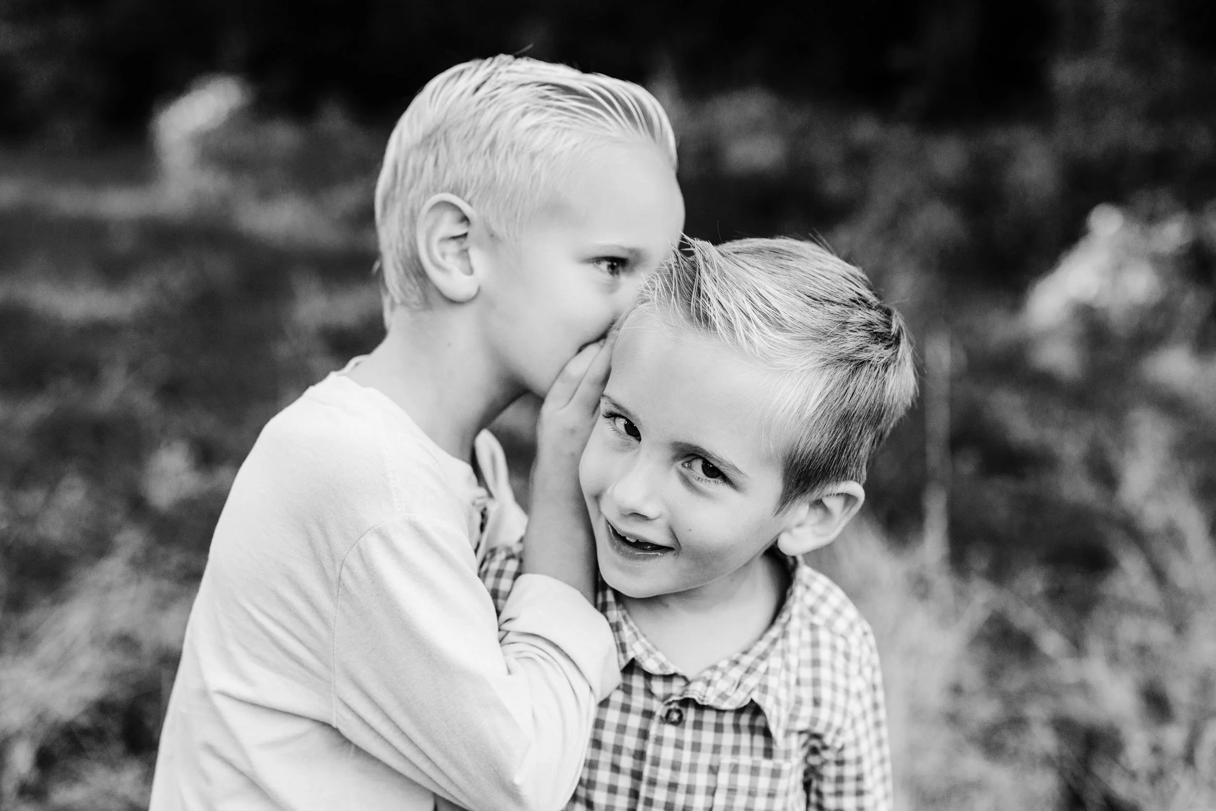 Two young boys with blonde hair sharing a secret outdoors in black and white
