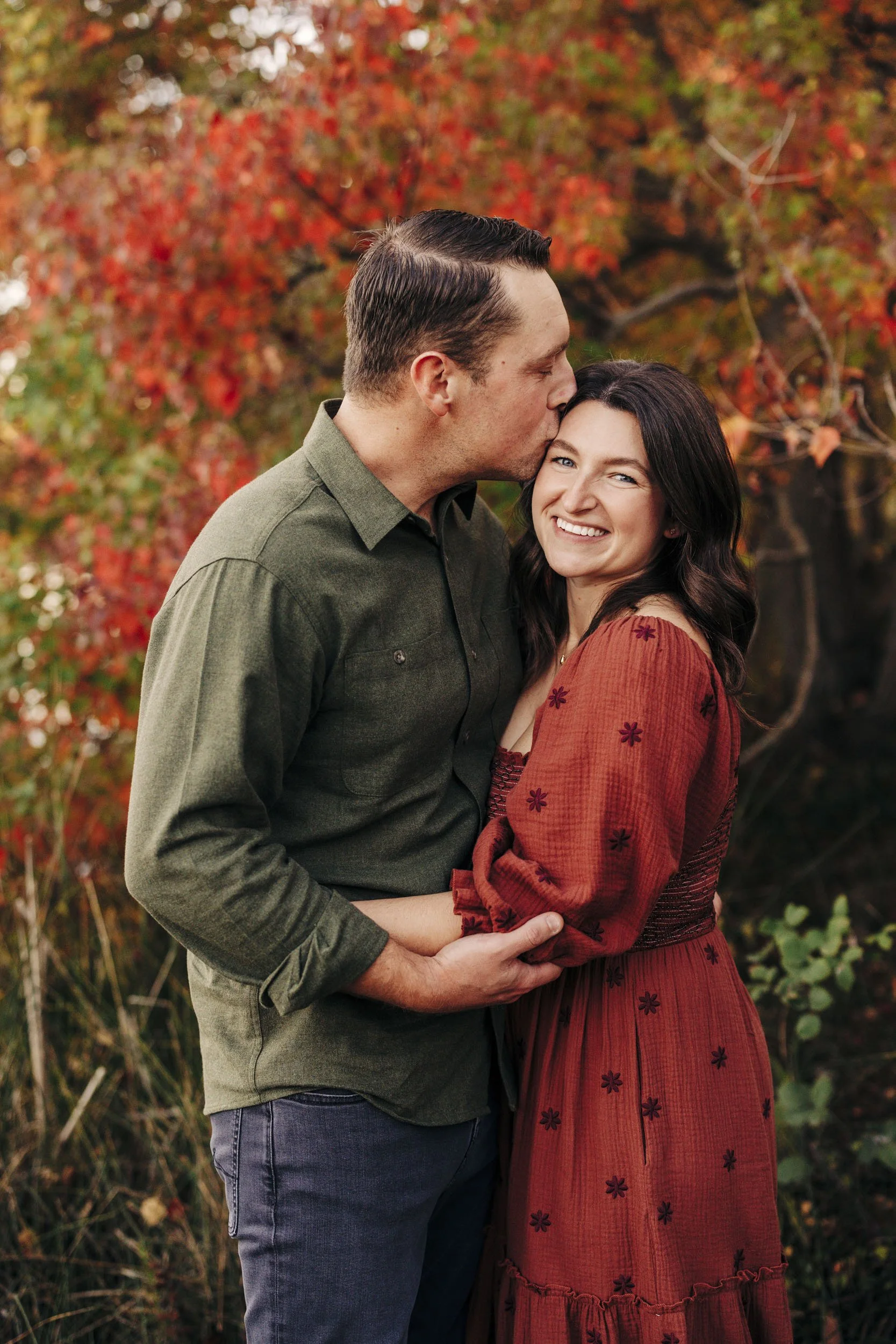 A couple standing outdoors in front of autumn trees. The man is kissing the woman's forehead while she smiles at the camera. The woman is wearing a red dress with embroidered flowers, and the man is wearing a green shirt and jeans.