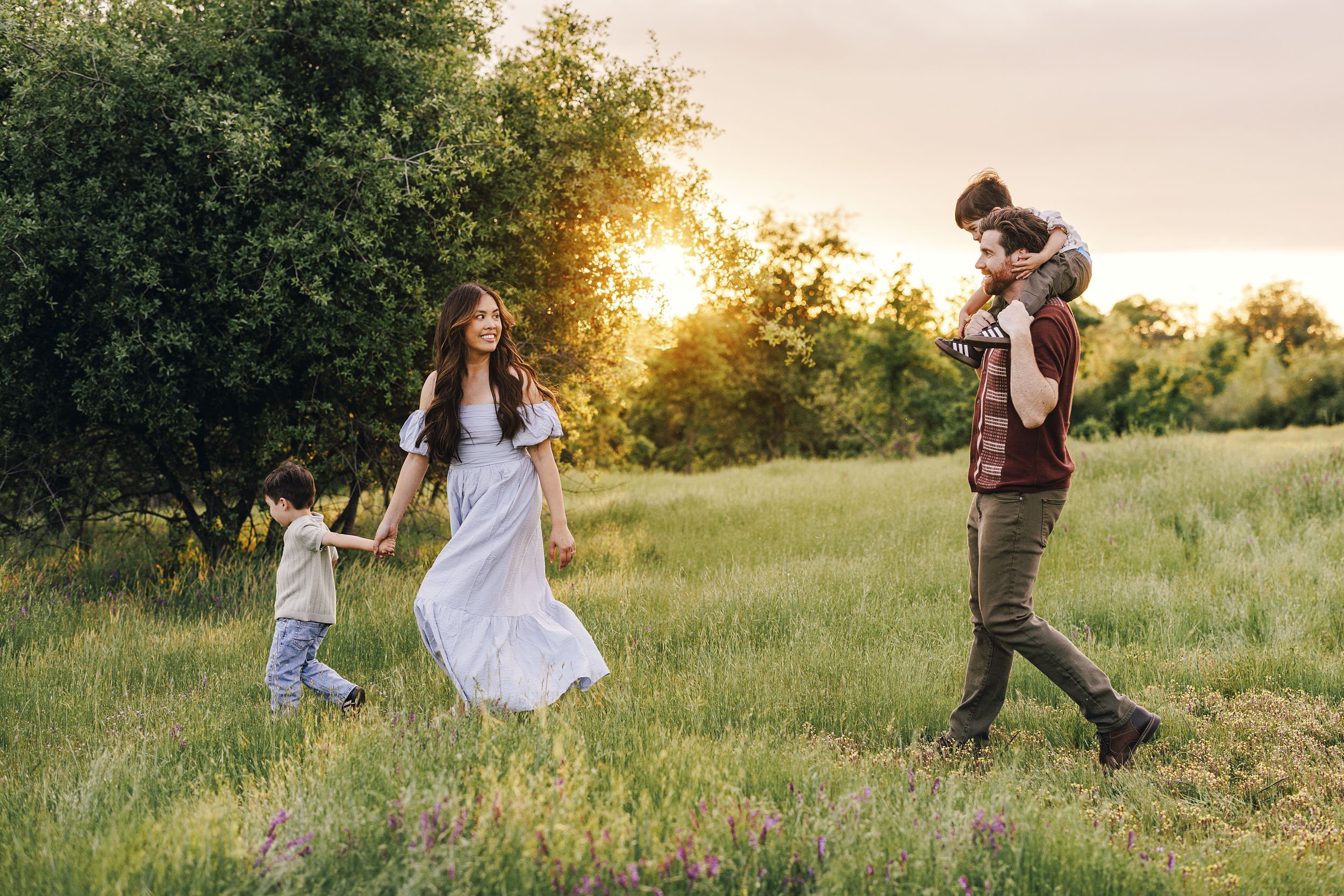 Woman in a white dress standing barefoot in a grassy field during sunset, holding her pregnant belly with both hands and looking down.