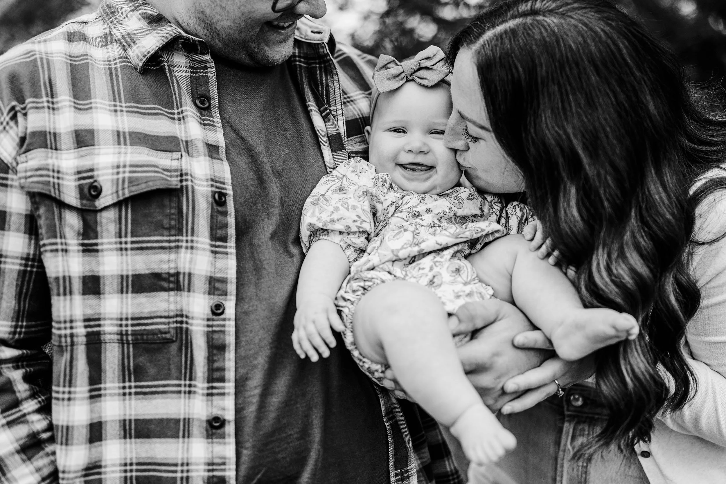 A happy baby being held and kissed by her mother, with a man, possibly her father, looking on outside. The baby is smiling, wearing a patterned dress and a bow in her hair.