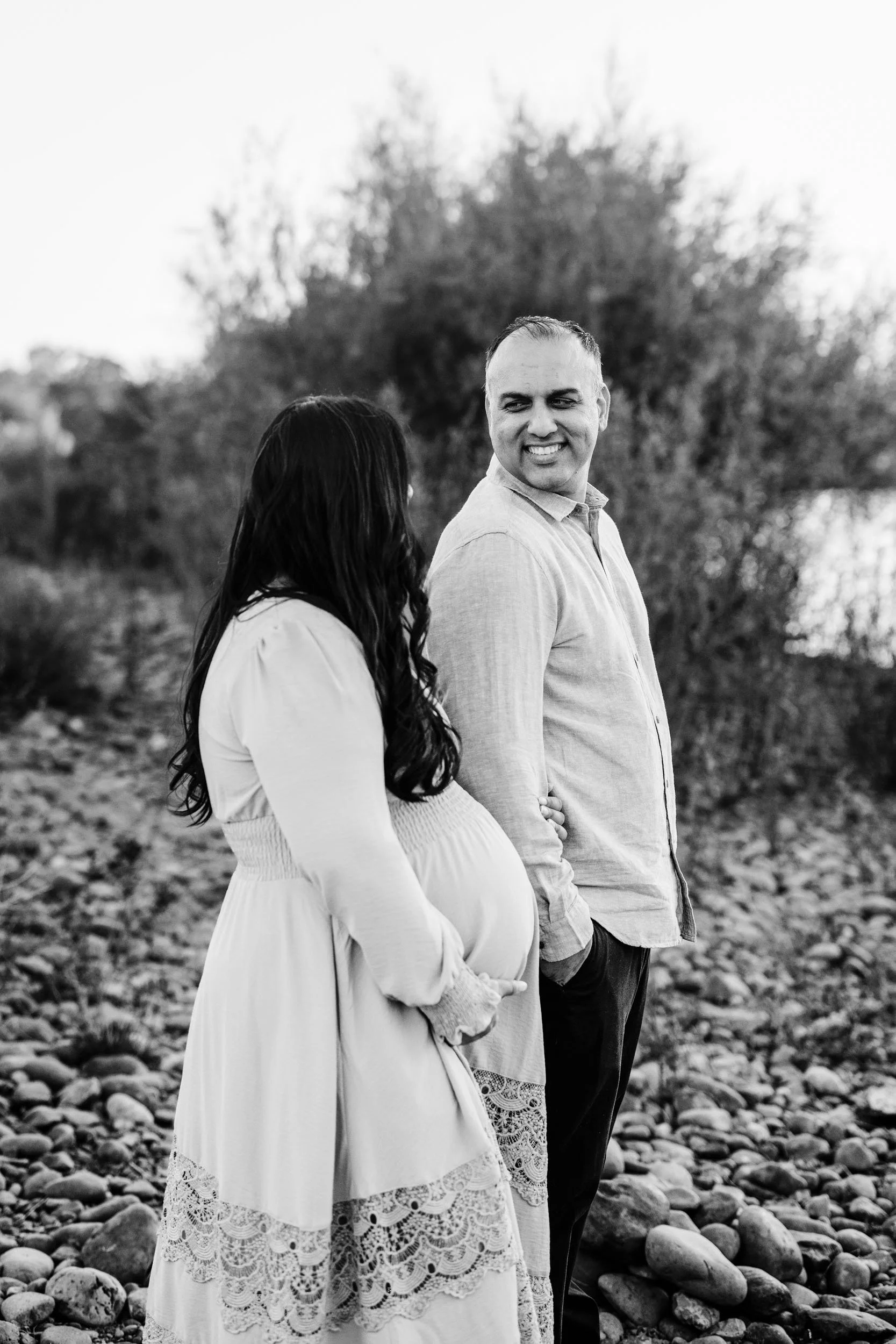 A pregnant woman with long dark hair wearing a white dress with lace details, and a man with short hair and a light-colored shirt, stand close outdoors on a rocky surface with trees in the background, smiling at each other.