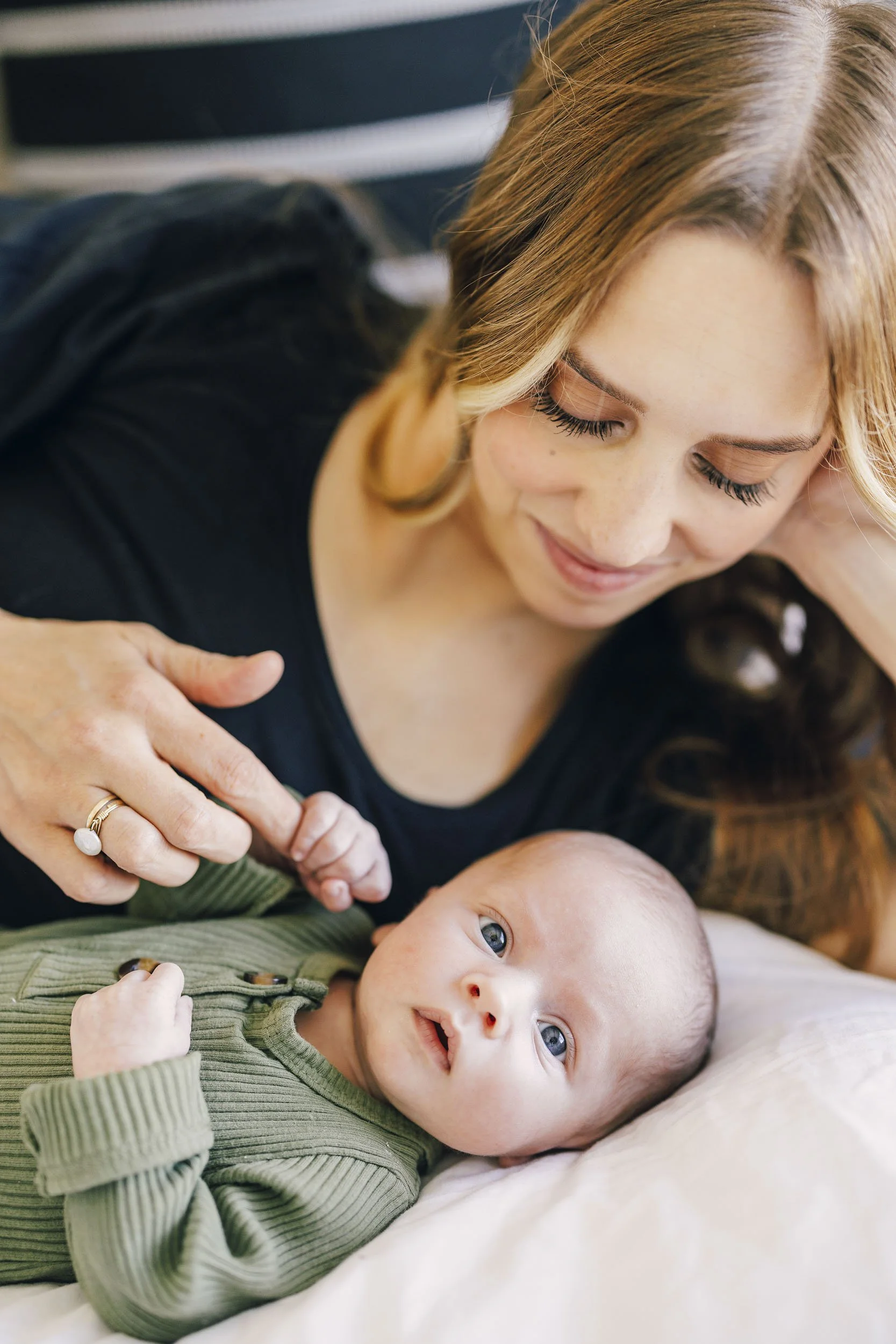 A woman with long red hair, smiling and lying next to a baby with blue eyes, on a bed, with the woman gently touching the baby's hand.