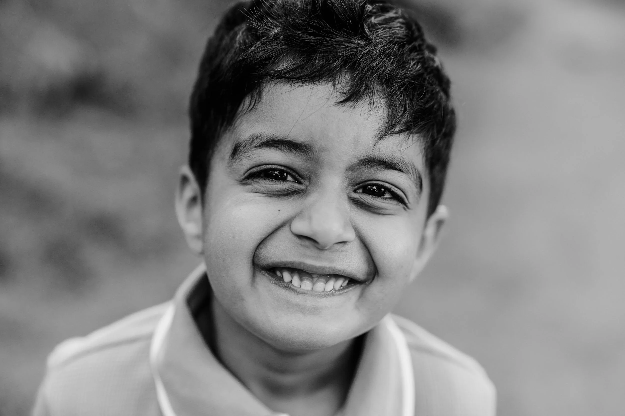 A young boy smiling with his teeth showing, in black and white, outside with blurred background.