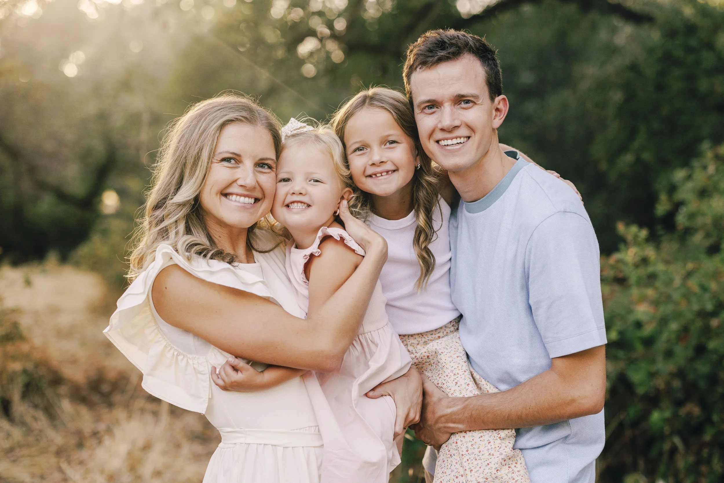 A happy family of four outdoors during sunset, smiling at the camera. The mother and father hold their two young daughters, all dressed in light-colored casual clothes during a family photo session with Amy Wright Photography.