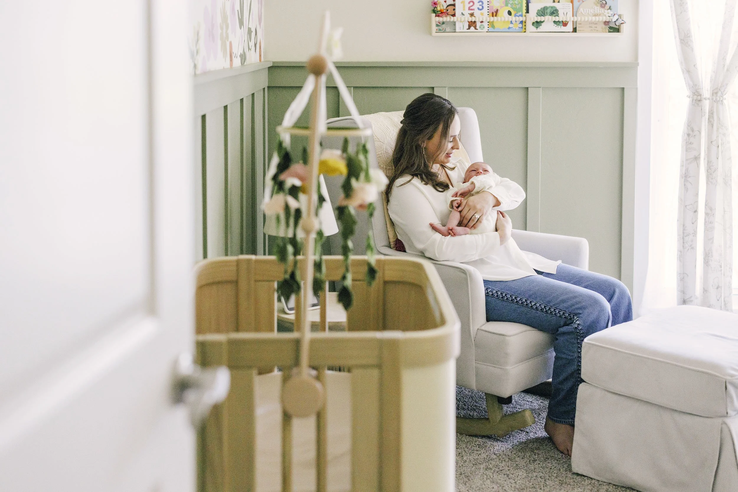 A woman sitting in a white armchair holding a baby in a bright room with green-paneled walls and floral curtains, with a wooden crib in the foreground.