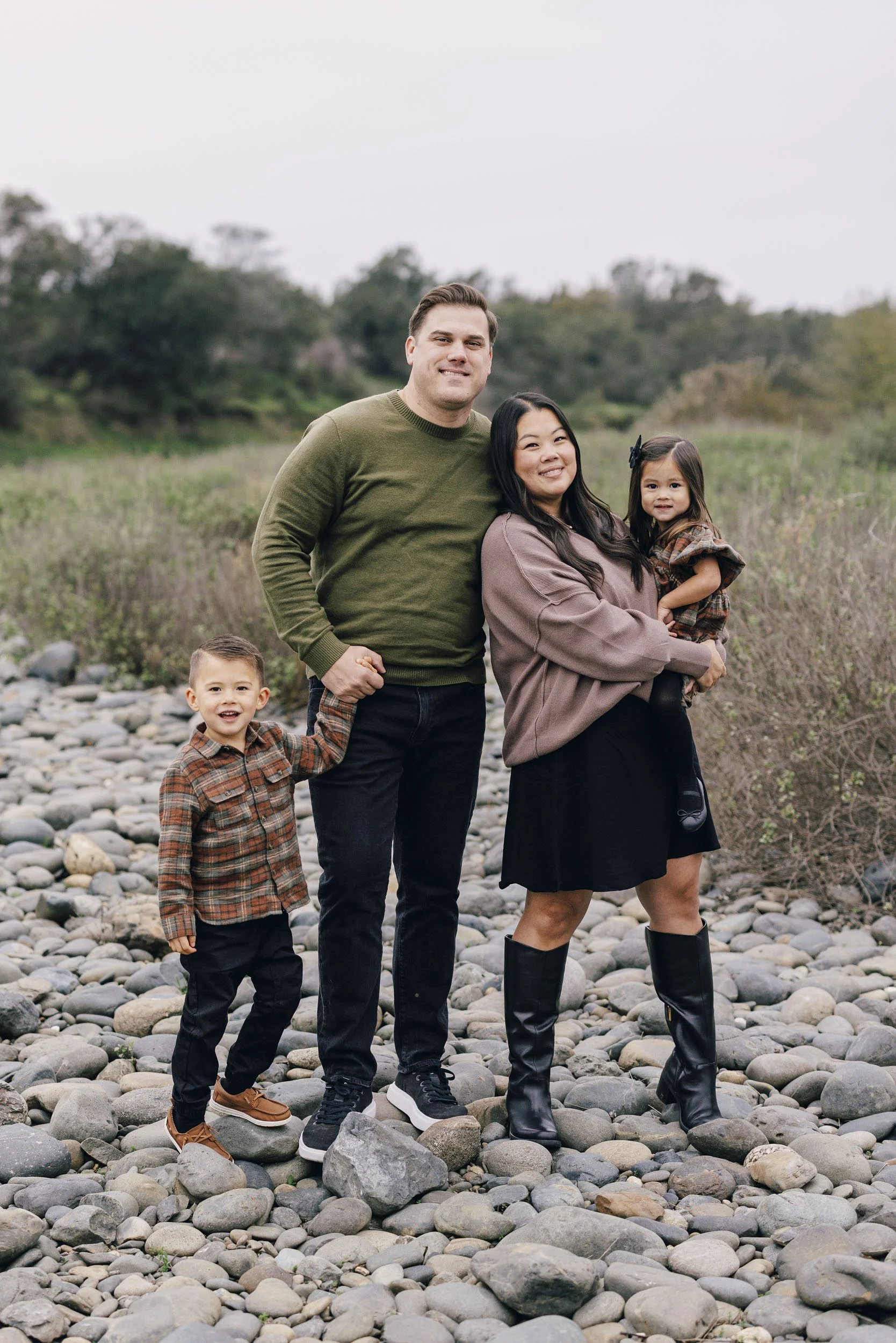 A family of four standing outdoors on a rocky riverbed with trees and bushes in the background, smiling at the camera.