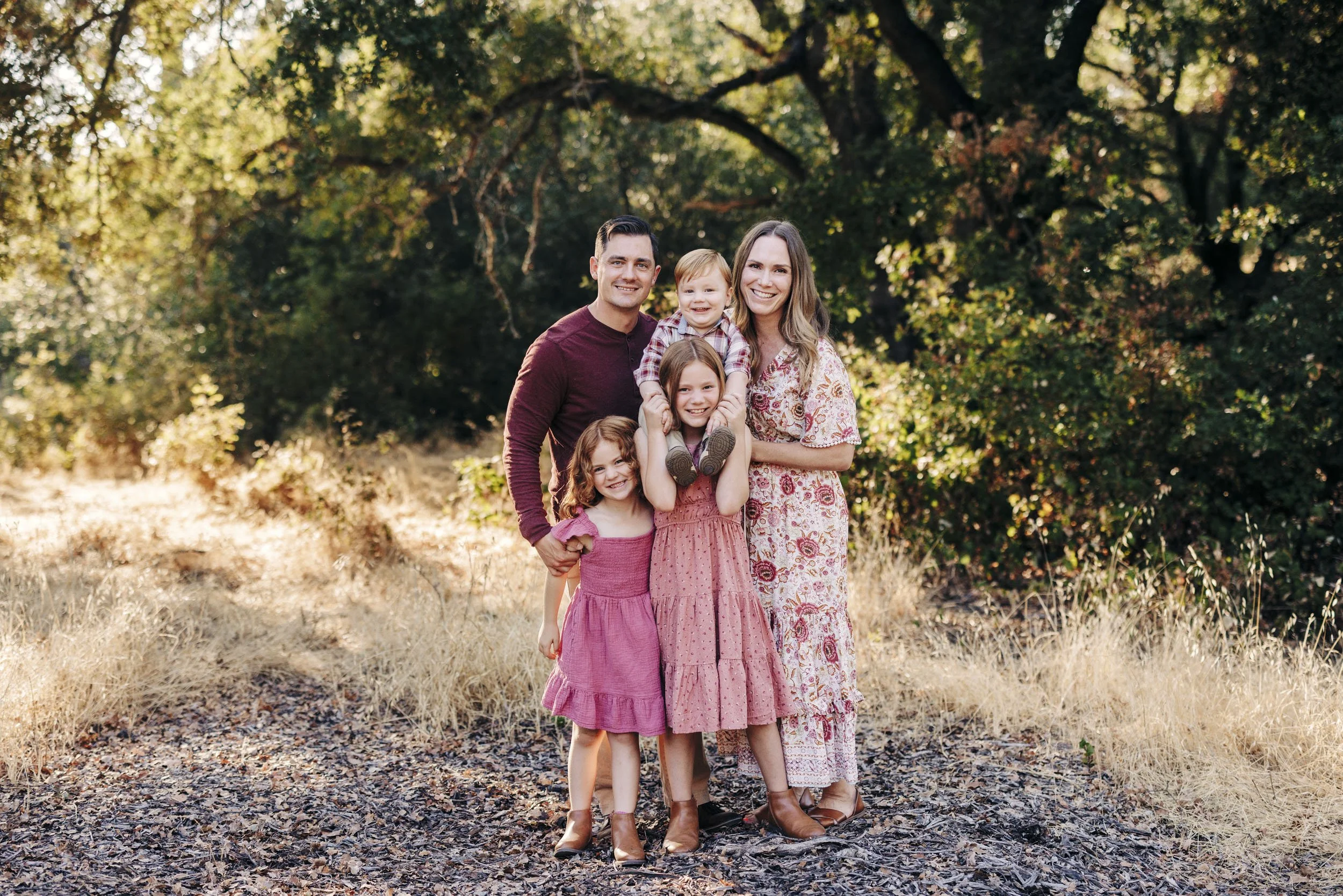 A family of five standing outdoors on a wooded trail, smiling at the camera. The family includes a man, woman, and three young girls, all dressed in casual fall clothing.