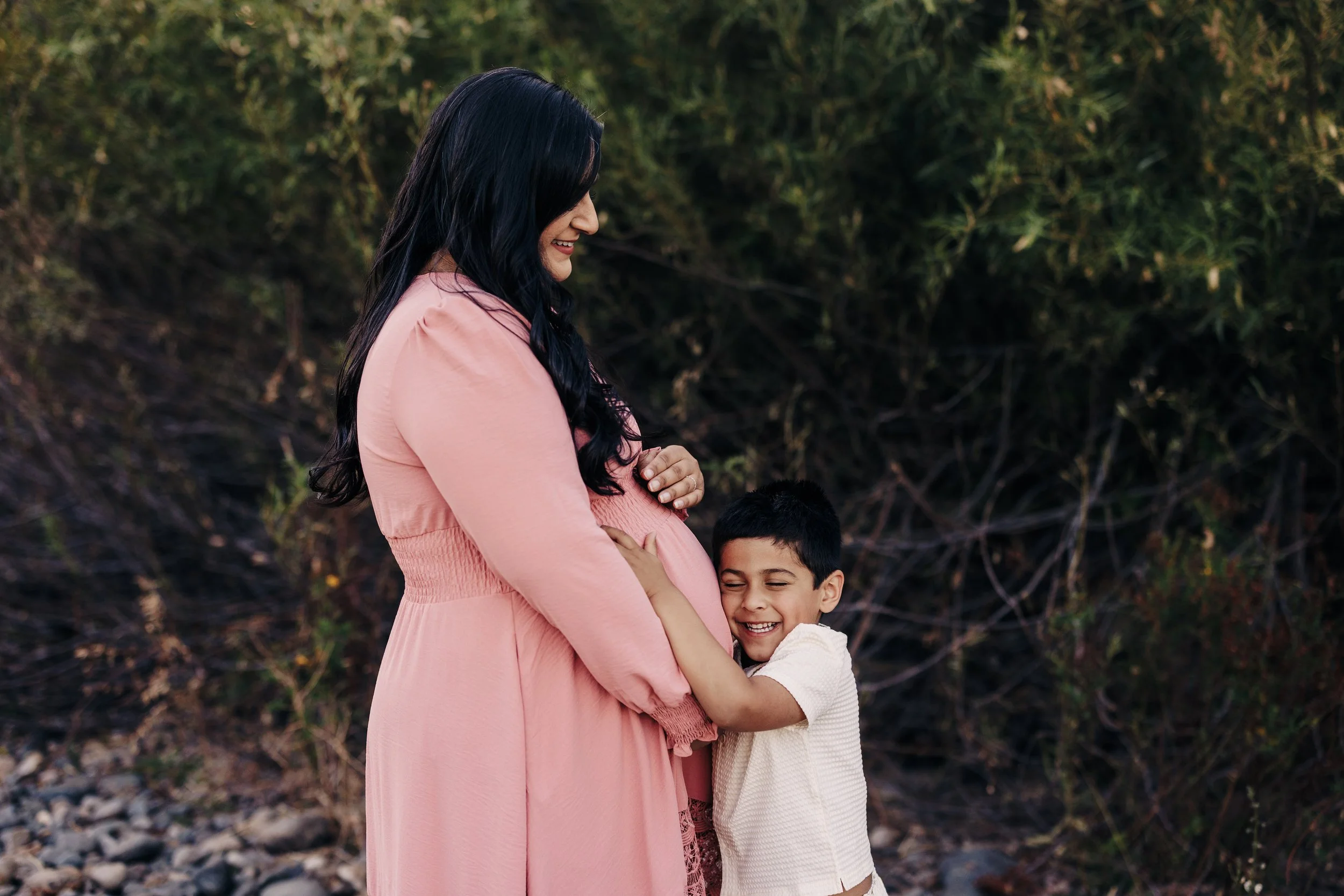 A pregnant woman in a pink dress smiling as a young boy hugs her belly outdoors with trees in the background.