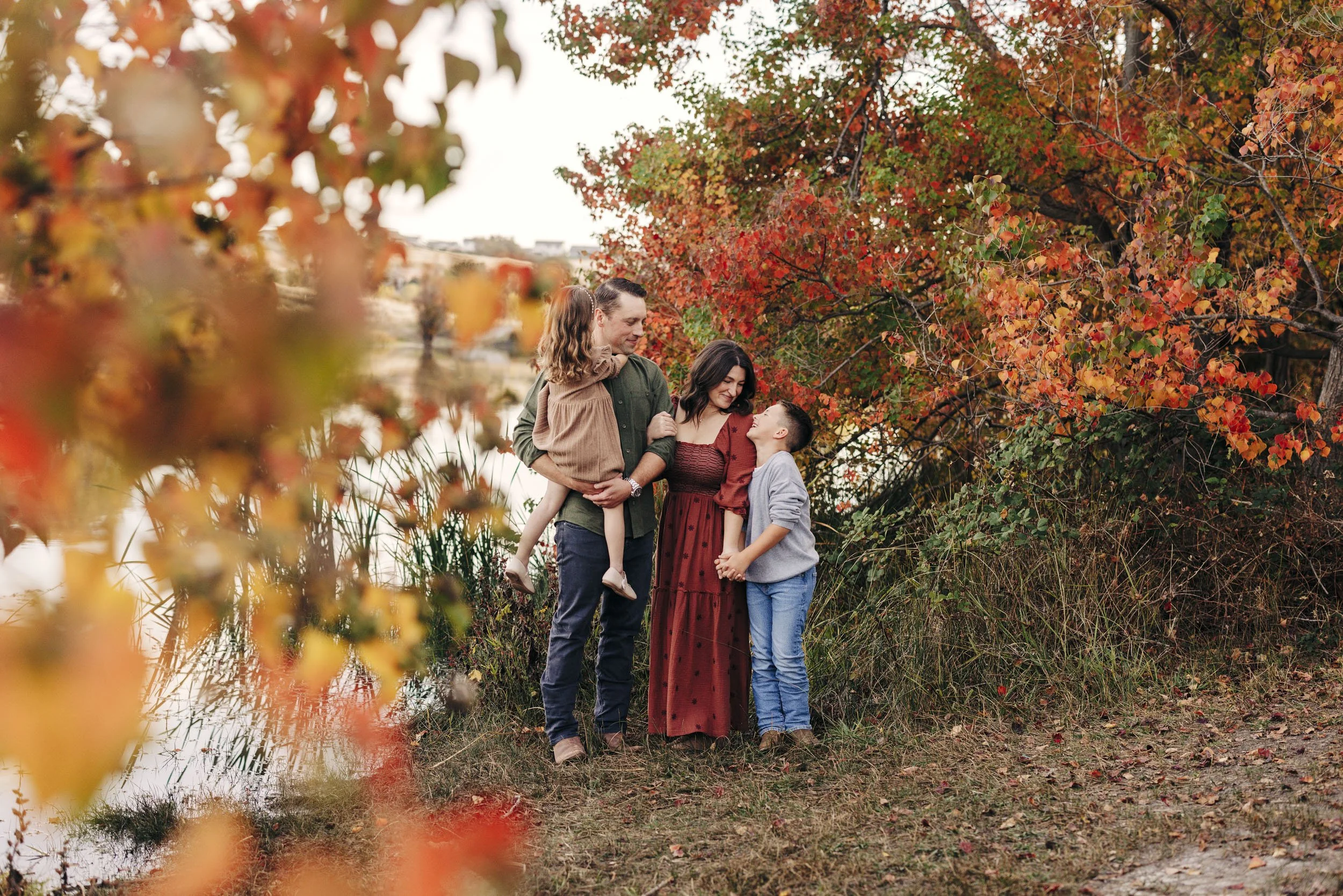 A family of four gets pictures taken outdoors near a lake, surrounded by vibrant fall trees with red, orange, and yellow leaves during the fall time in Lincoln, California.