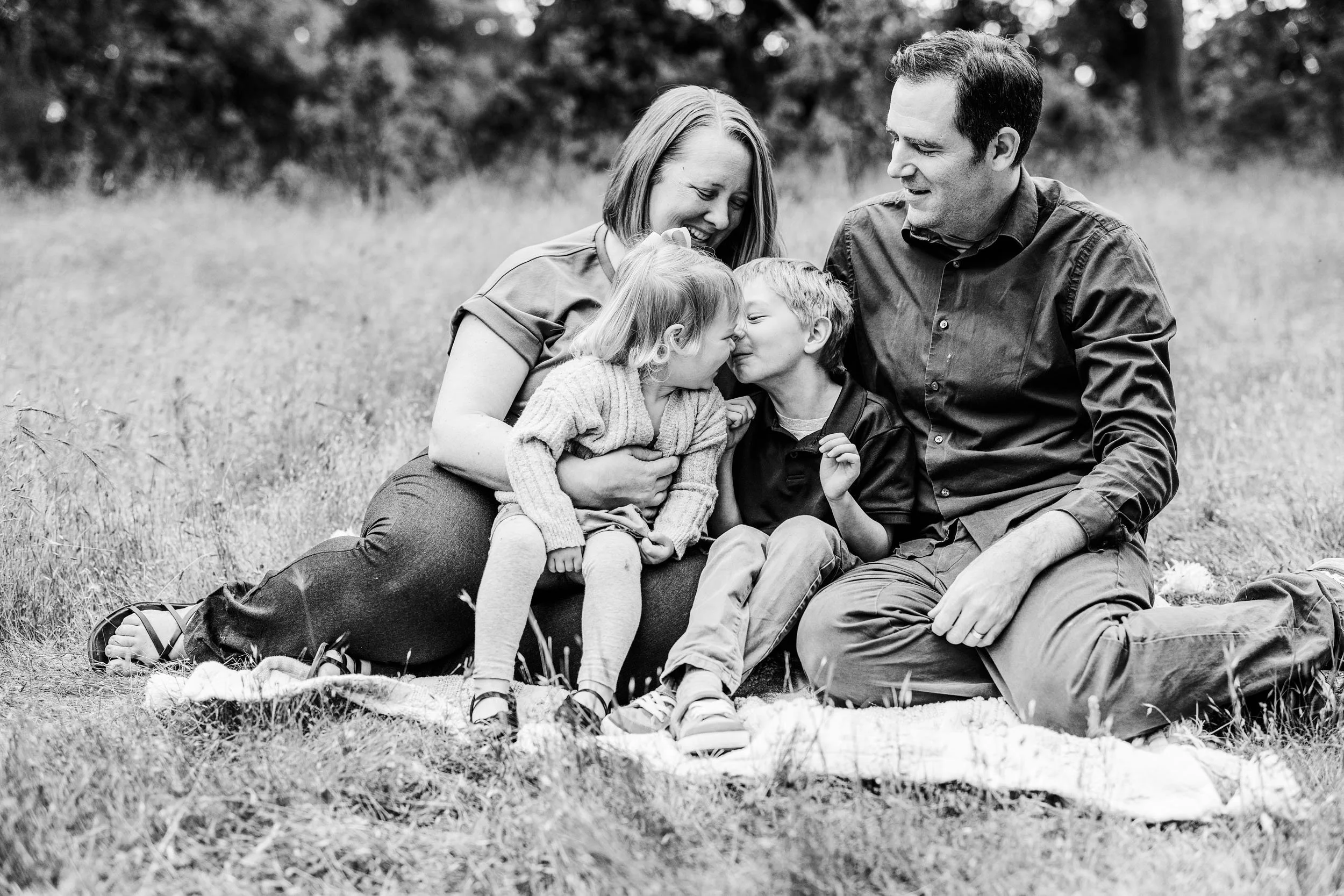 A black and white photo of a family sitting on a blanket outdoors in a grassy field, with a mother, father, and two young children touching noses in a playful, affectionate moment.