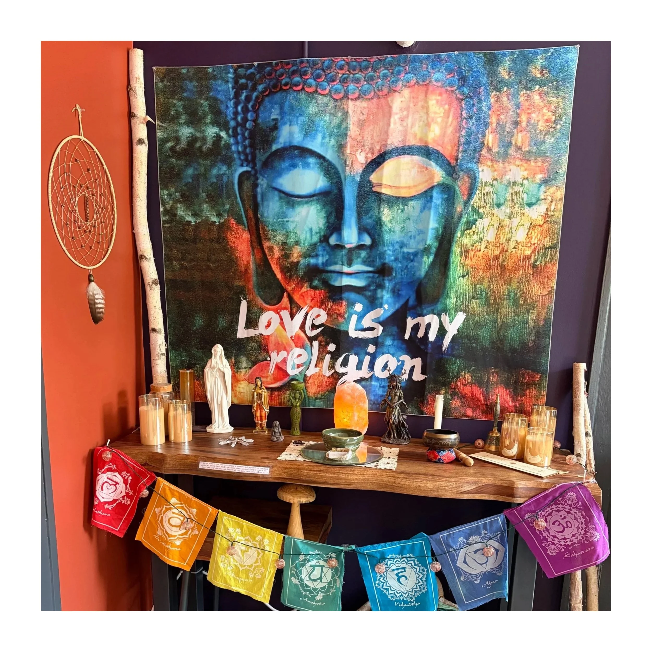 A space with a colorful tapestry and spiritual objects. The tapestry features a serene face with closed eyes, surrounded by vibrant colors and abstract patterns. Stable candles, small statues, and colorful prayer flags are arranged on the table below
