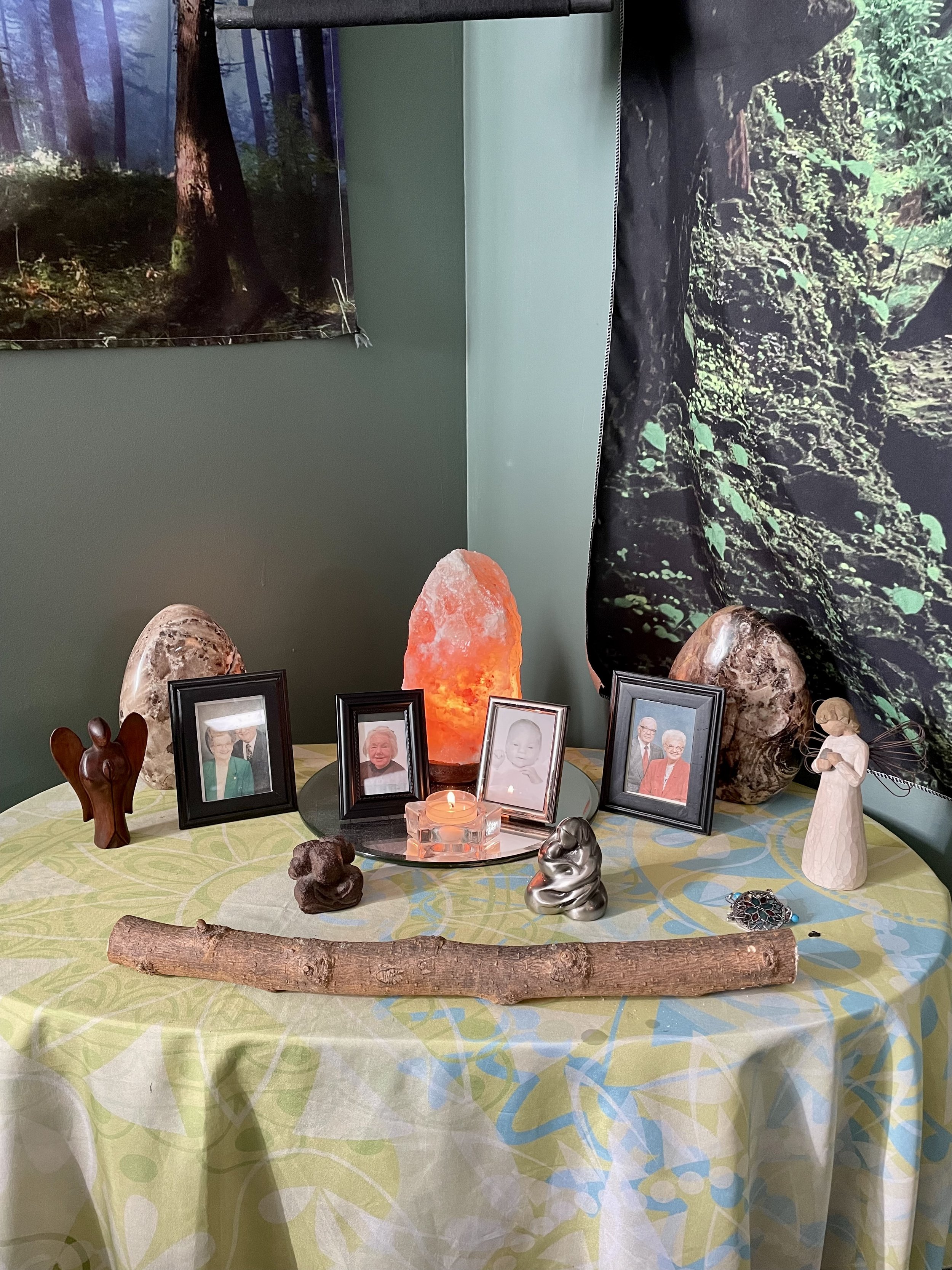 Home altar with framed photos, salt lamp, wooden stick, candles, and figurines on a round table with a patterned cloth.