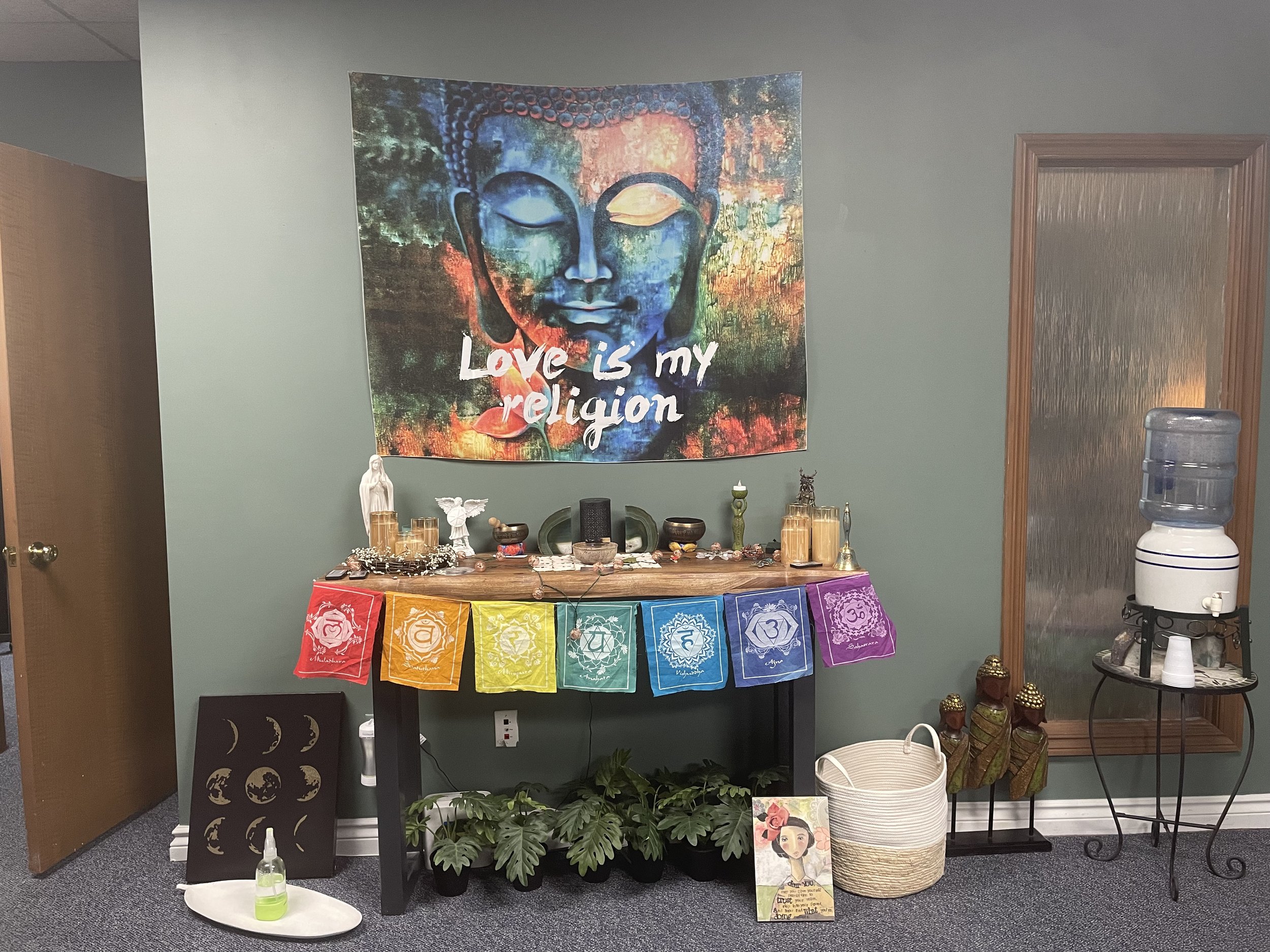 A meditation altar with a tapestry of a Buddha face with the phrase 'Love is my religion.' The altar is decorated with prayer flags in chakra colors, candles, statues, crystals, and incense. A water cooler is on the right, and greenery and artwork ar
